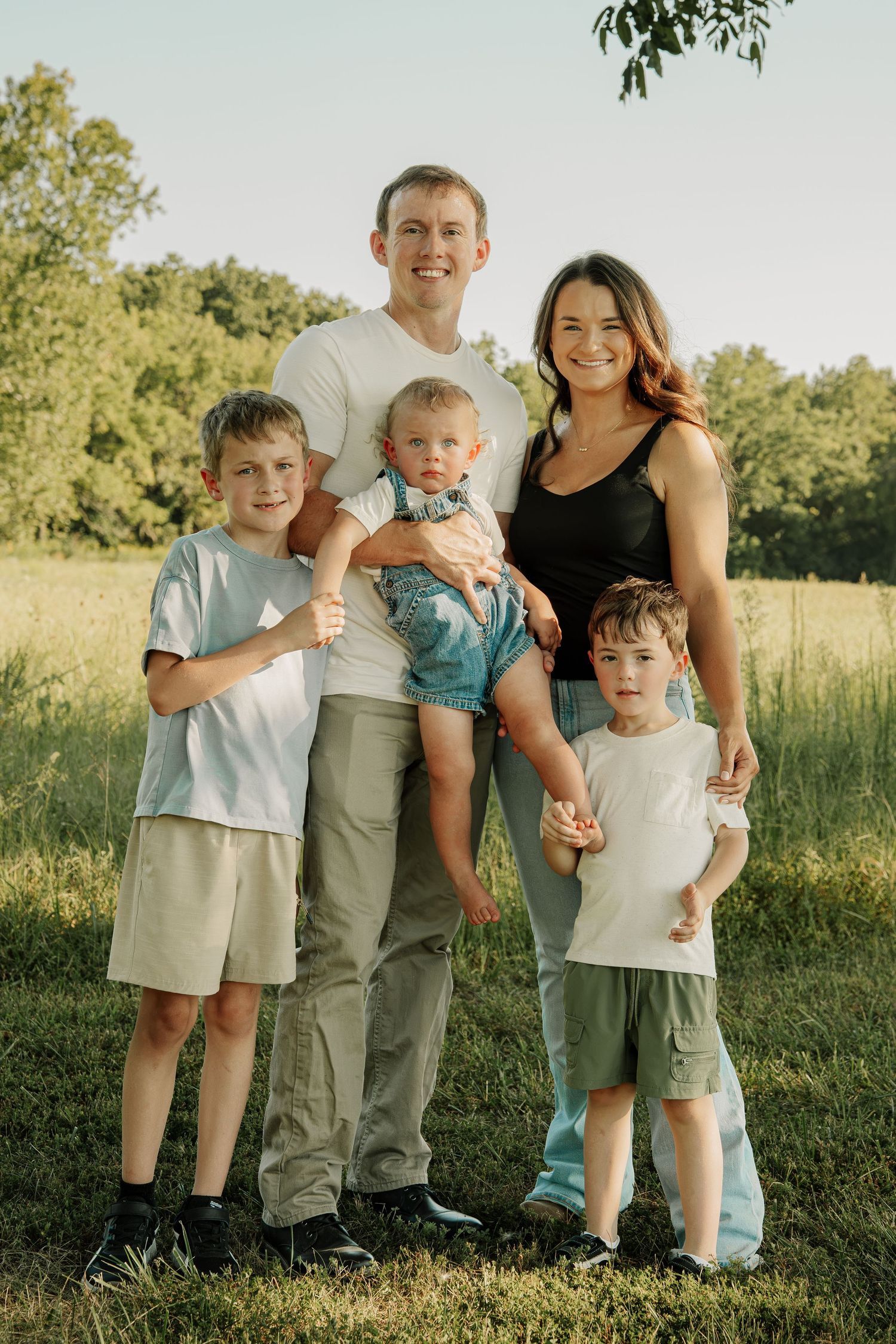 Family of five posing outdoors in a green field, parents with three young boys smiling together on a sunny day.