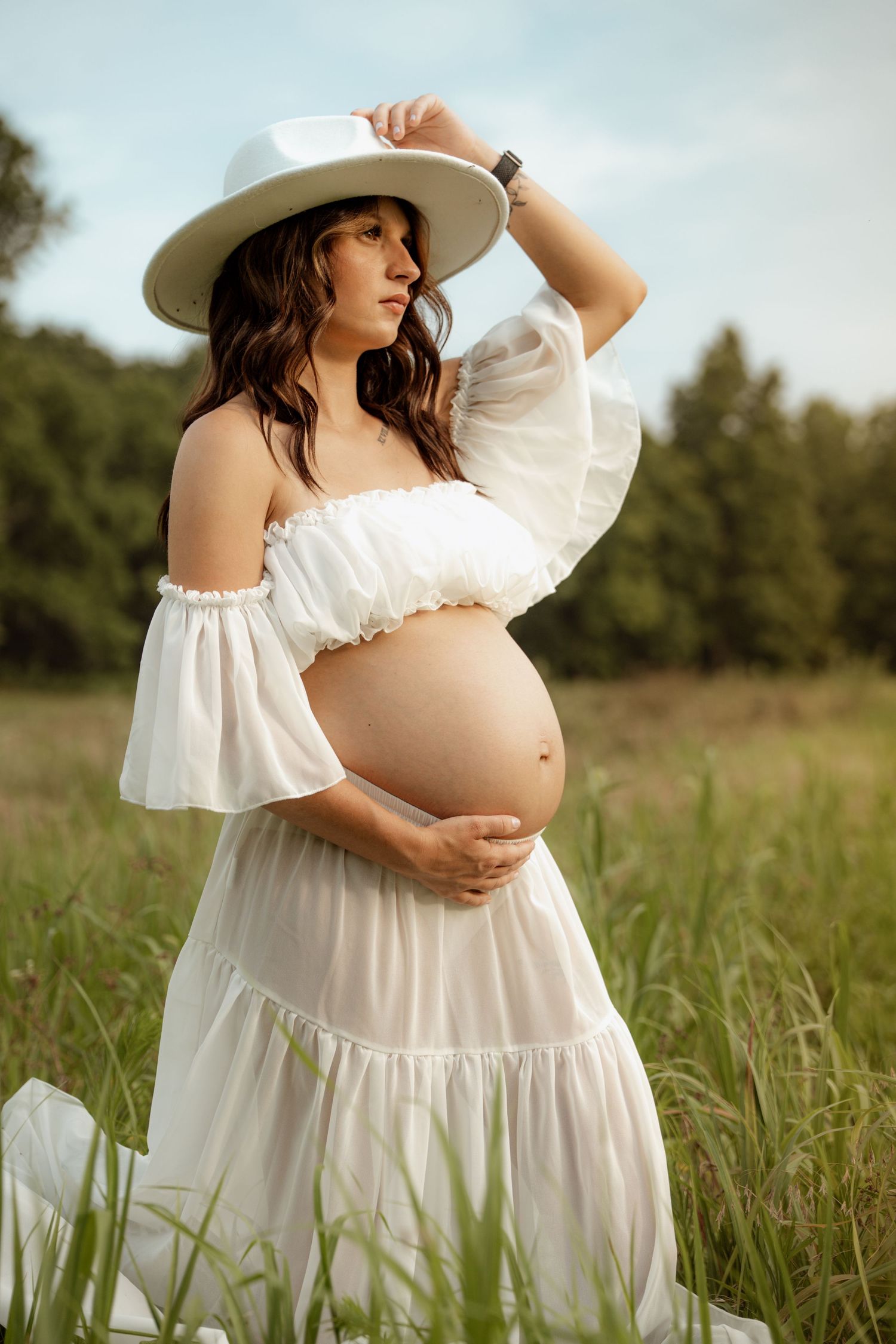 Pregnant woman in white off-shoulder crop top and skirt holds belly while touching wide-brim hat in green field.