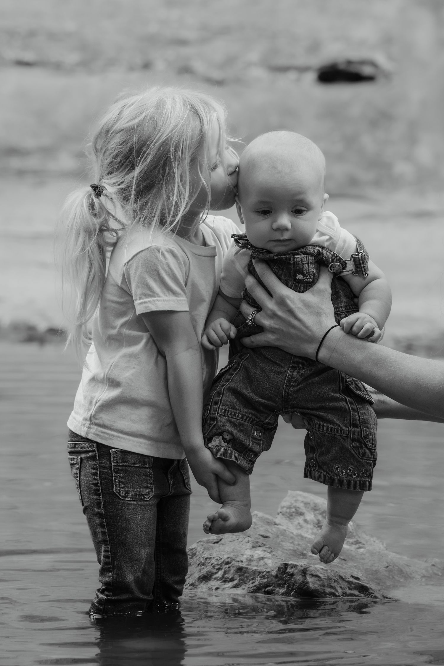 Black and white photo of a young girl kissing a baby held by an adult hand while standing in shallow water.