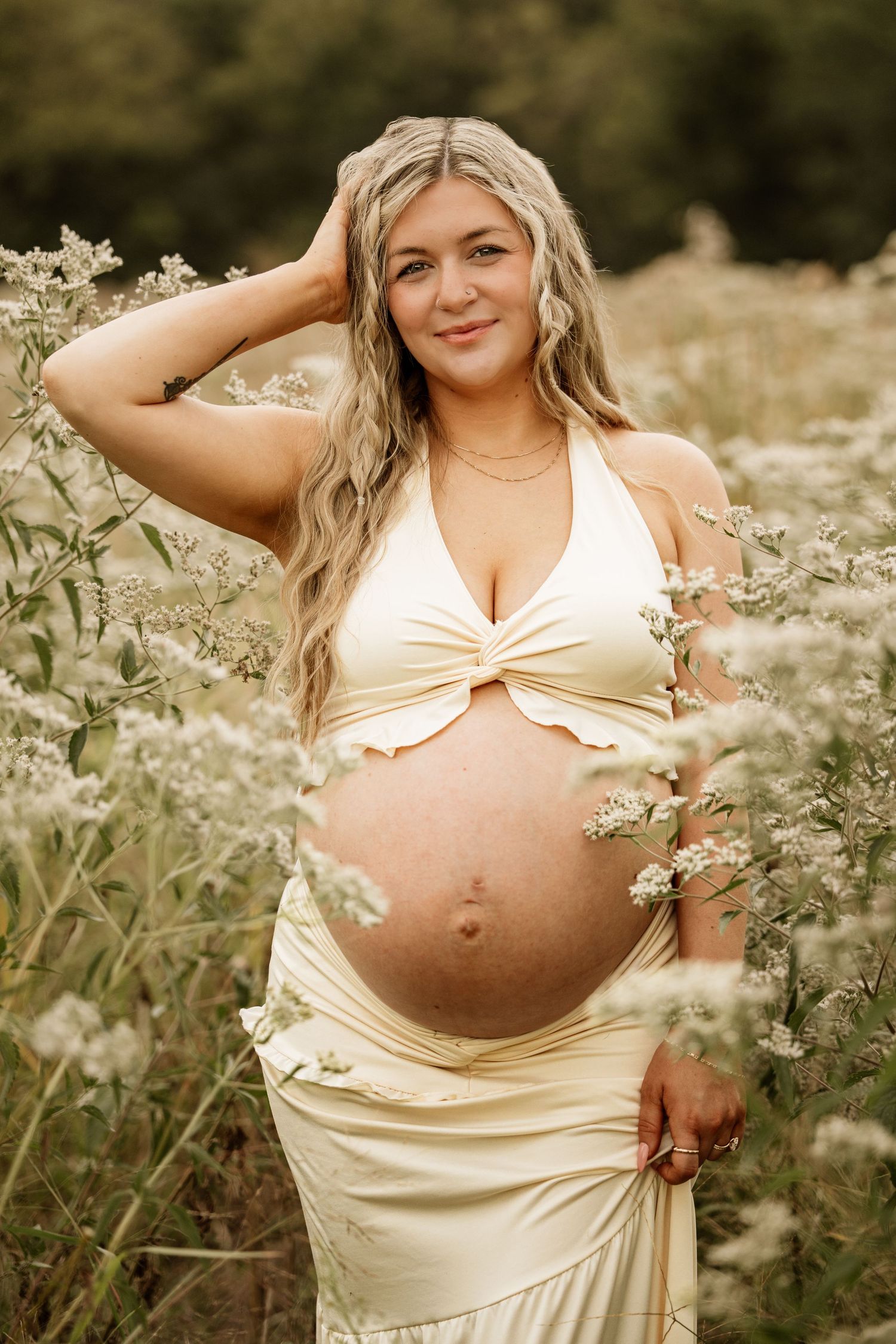 Smiling pregnant woman in cream halter dress standing among white wildflowers in a lush green field.