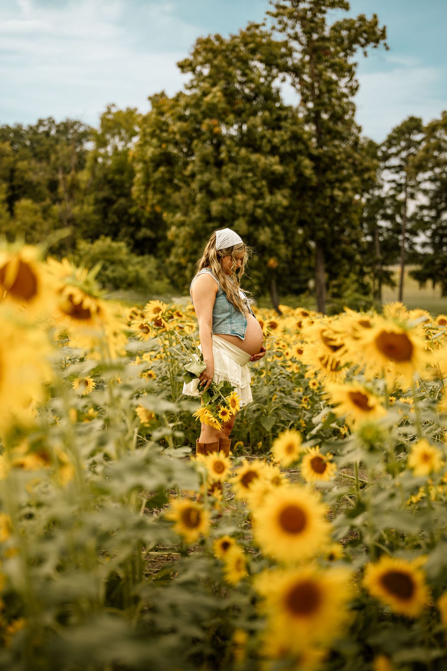 Pregnant woman in white headband and denim top stands in a vibrant sunflower field holding a bouquet.