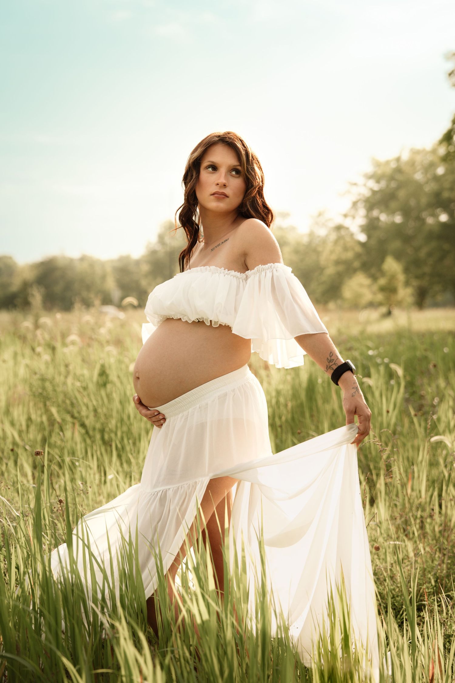 Pregnant woman in white boho dress holding her belly while standing in a sunlit green field.