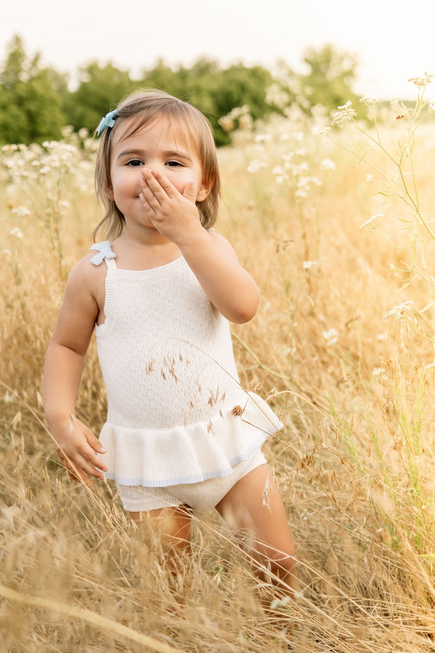 Cute toddler girl in white romper covering her mouth while standing in a sunny field.