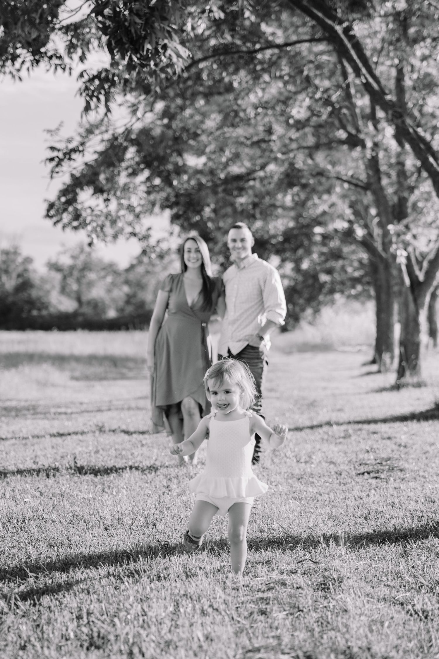 Black and white photo of a happy family with a toddler girl running in a sunny park with large trees.