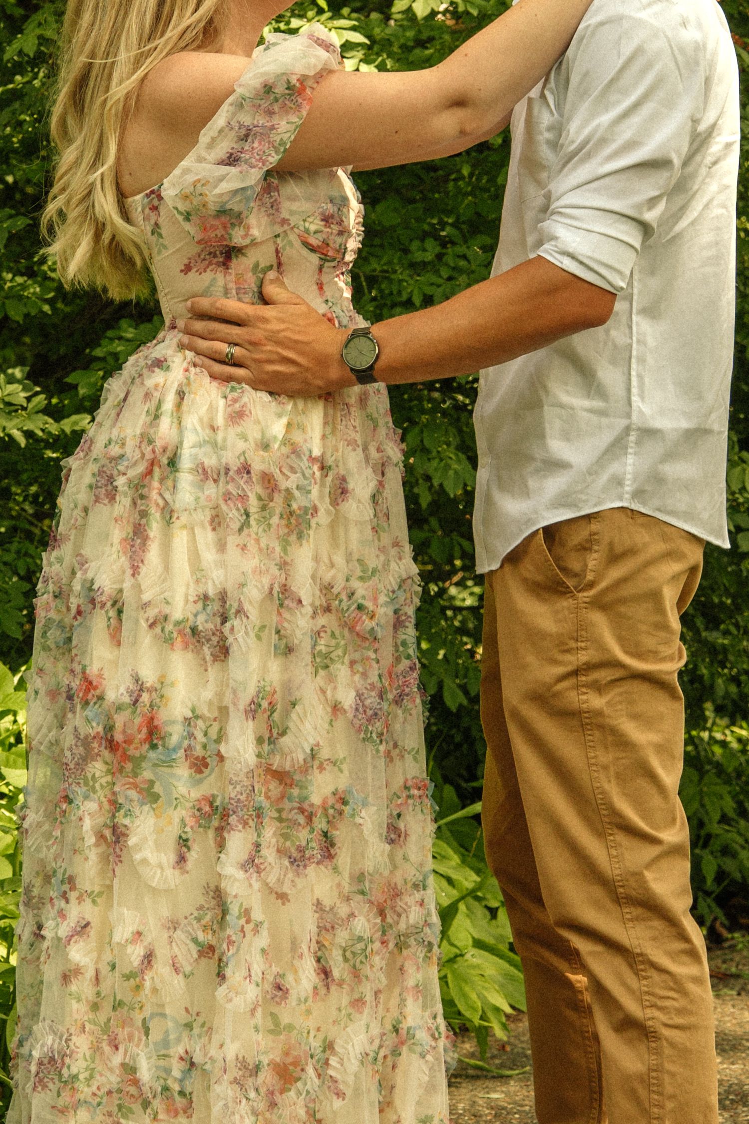 Couple embracing outdoors, woman in floral maxi dress, man in white shirt and tan pants, surrounded by lush greenery.