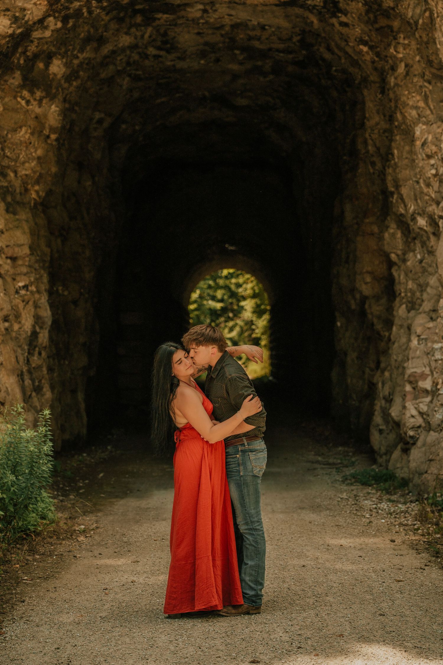 Couple embracing and kissing in a rustic stone tunnel, woman in red dress, man in jeans, romantic engagement photo.