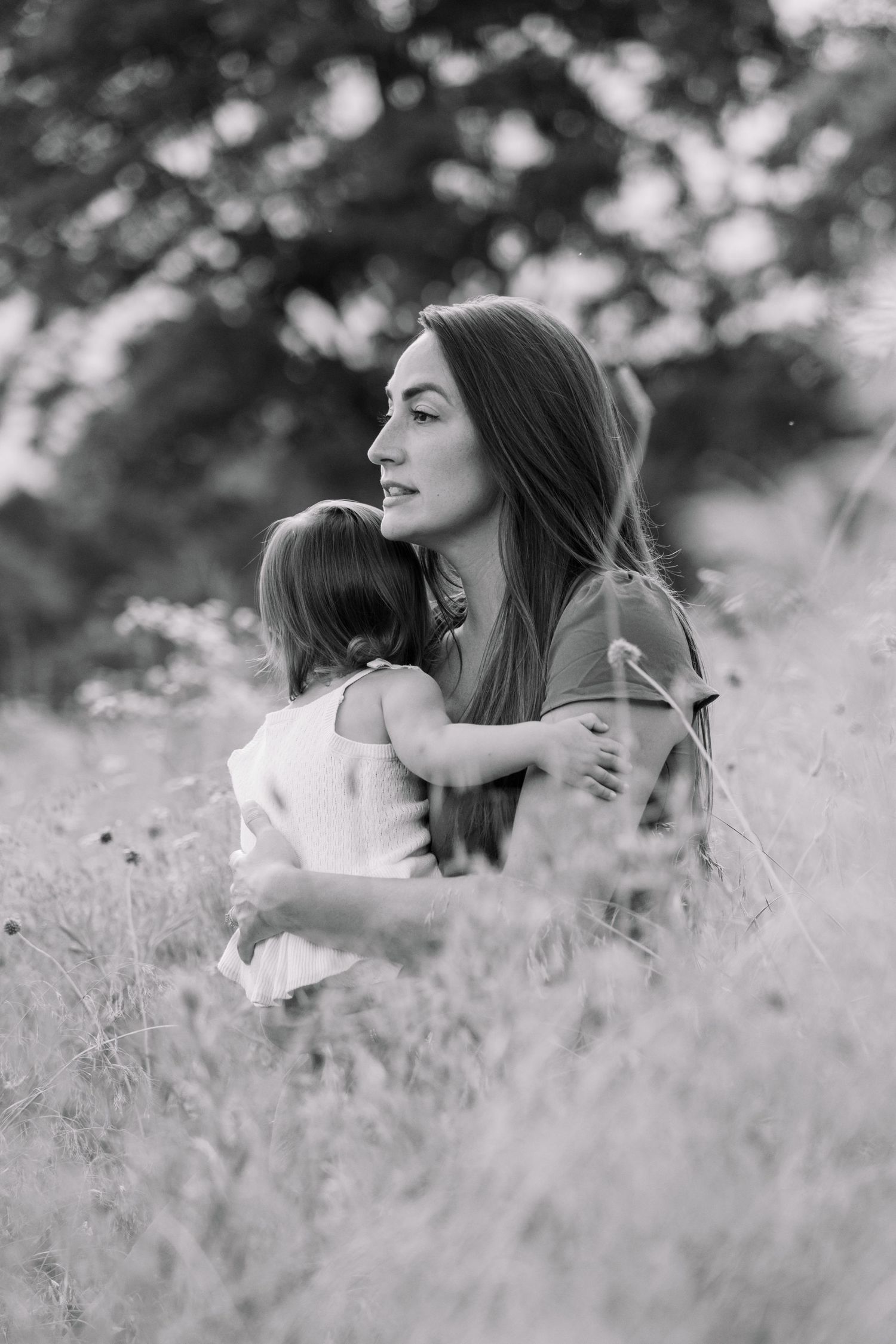 Black and white photo of a mother tenderly holding her young daughter in a field of tall grass with trees behind them.