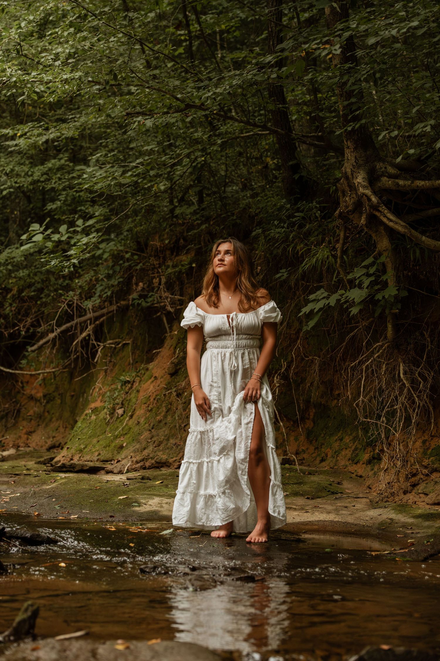 Woman in white off-shoulder dress standing barefoot in a serene forest creek.