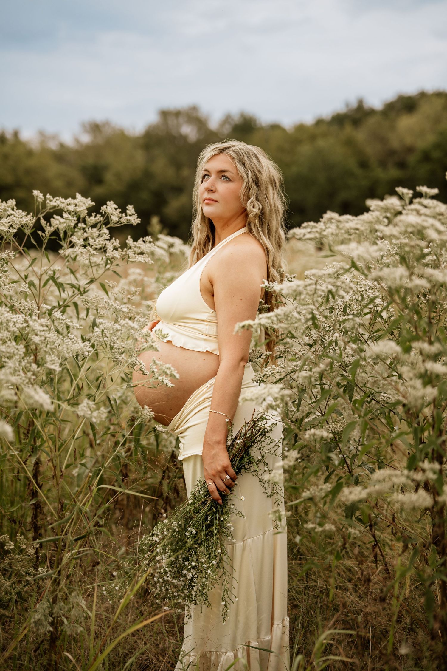 Pregnant woman in cream dress holding wildflowers in a lush field, maternity photography outdoors.