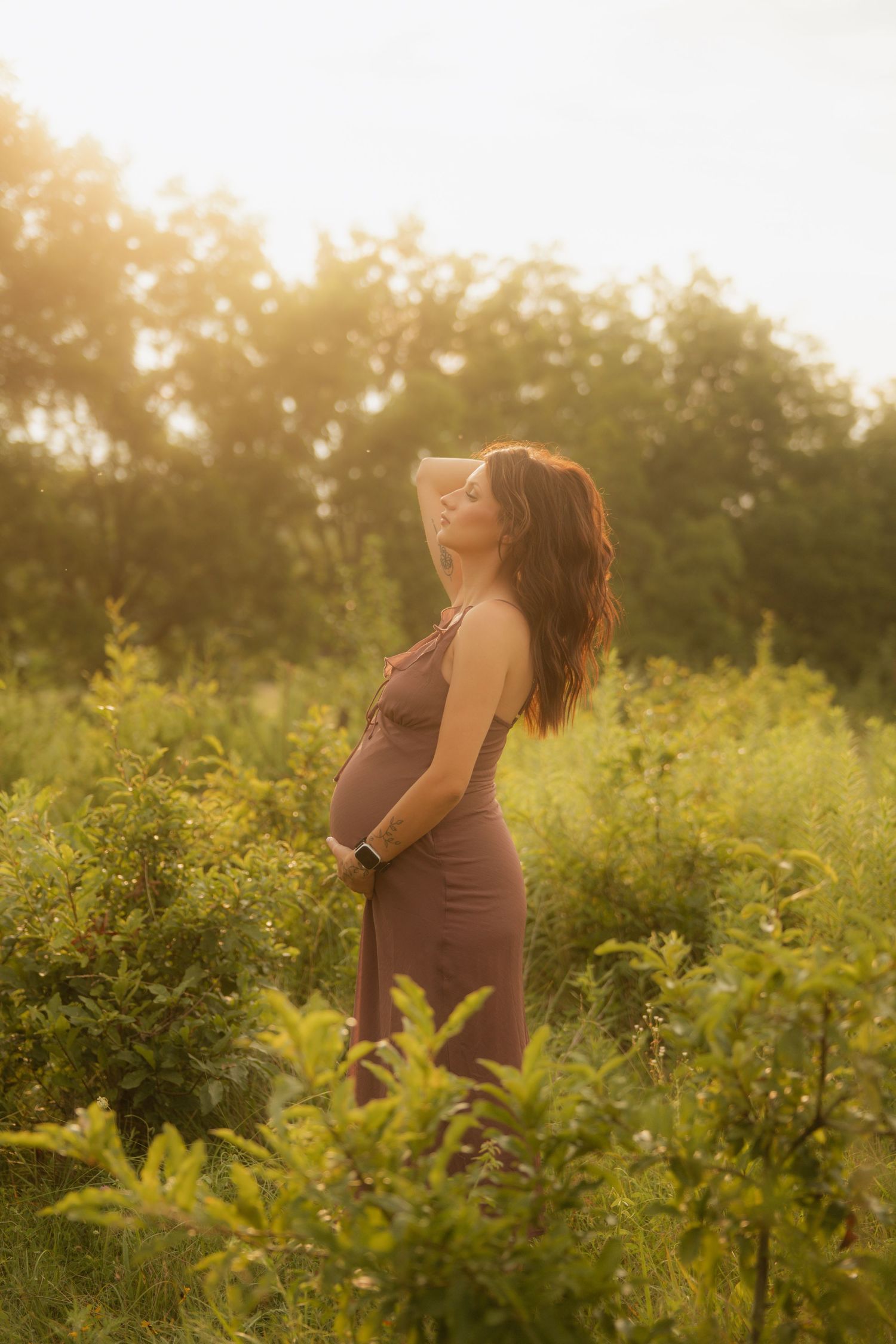 Pregnant woman in a brown dress standing in a sunlit green field during golden hour.