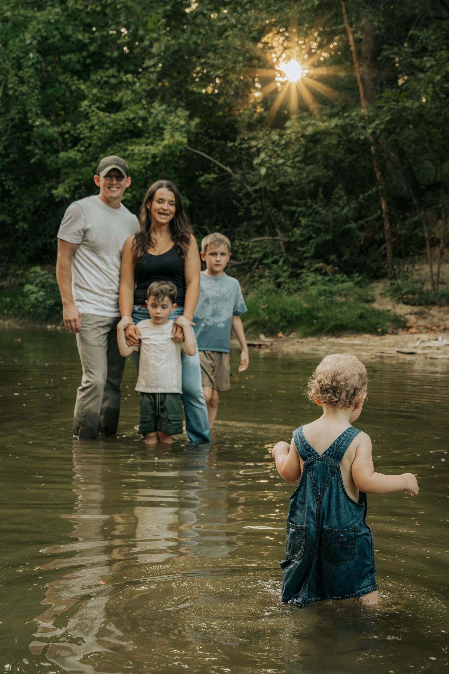 Family of five wading in shallow creek at sunset surrounded by lush forest trees.