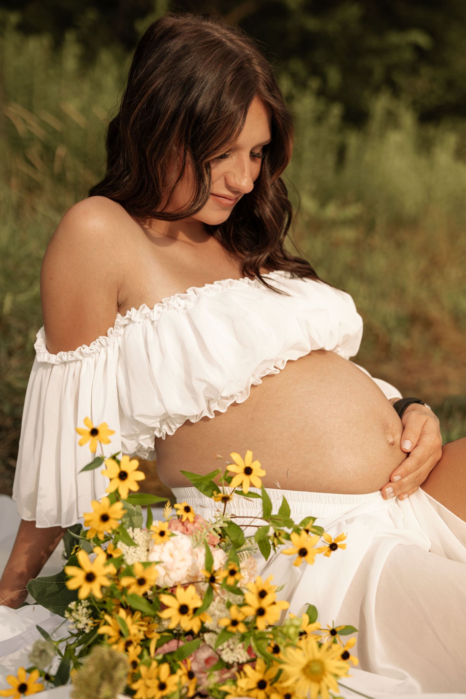 Pregnant woman in white off-shoulder dress gazes down at baby bump with yellow flowers in foreground.