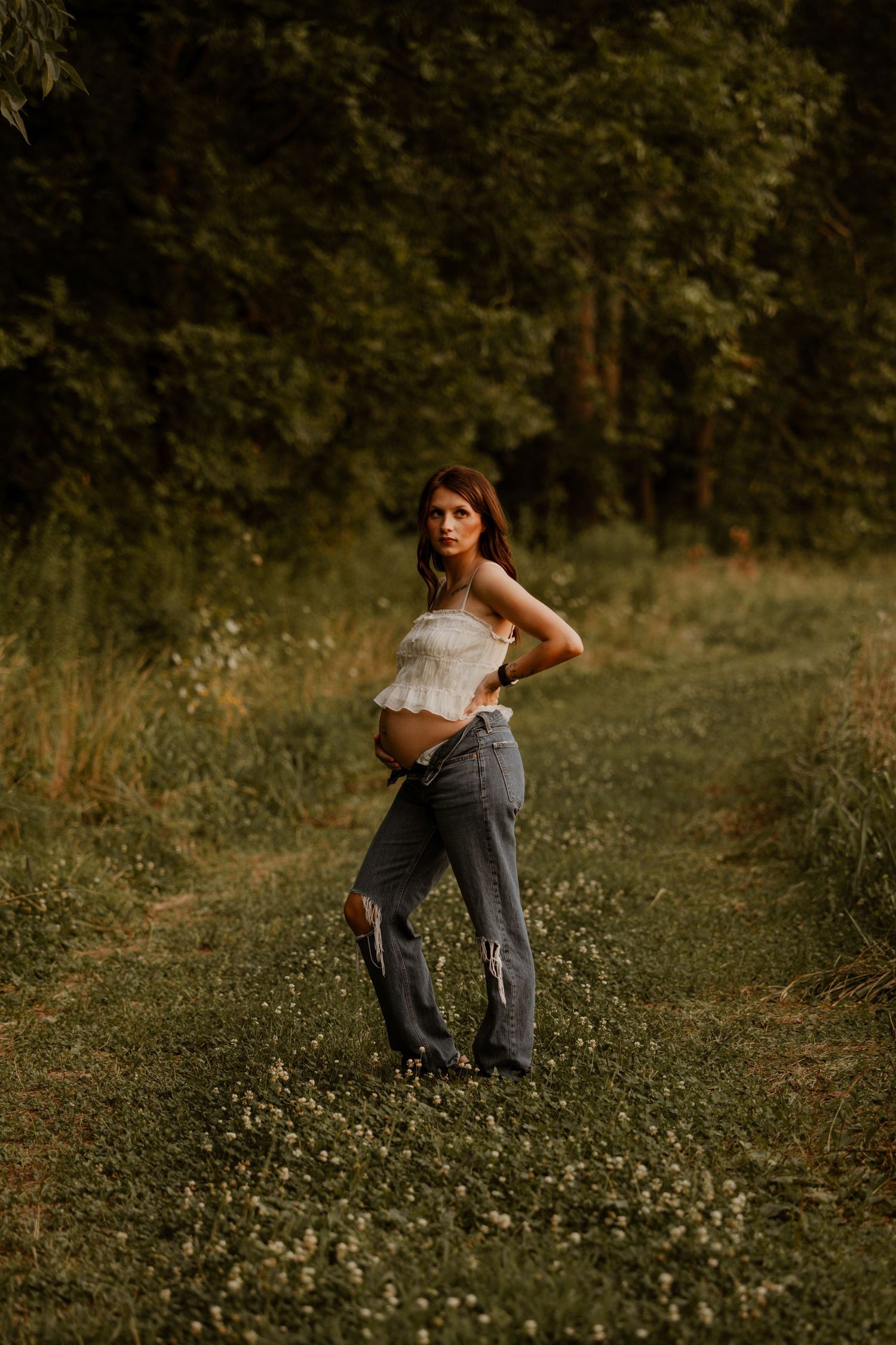 Pregnant woman in white crop top and ripped jeans poses on a grassy forest path during golden hour.
