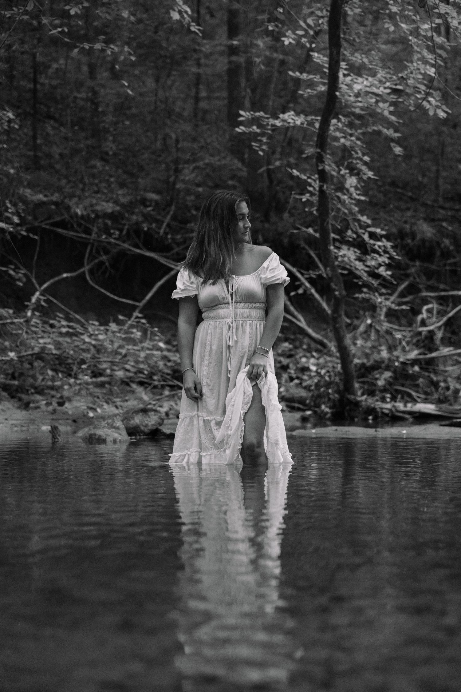 Black and white photo of a woman in a white lace dress standing in a forest stream, reflecting in the water.