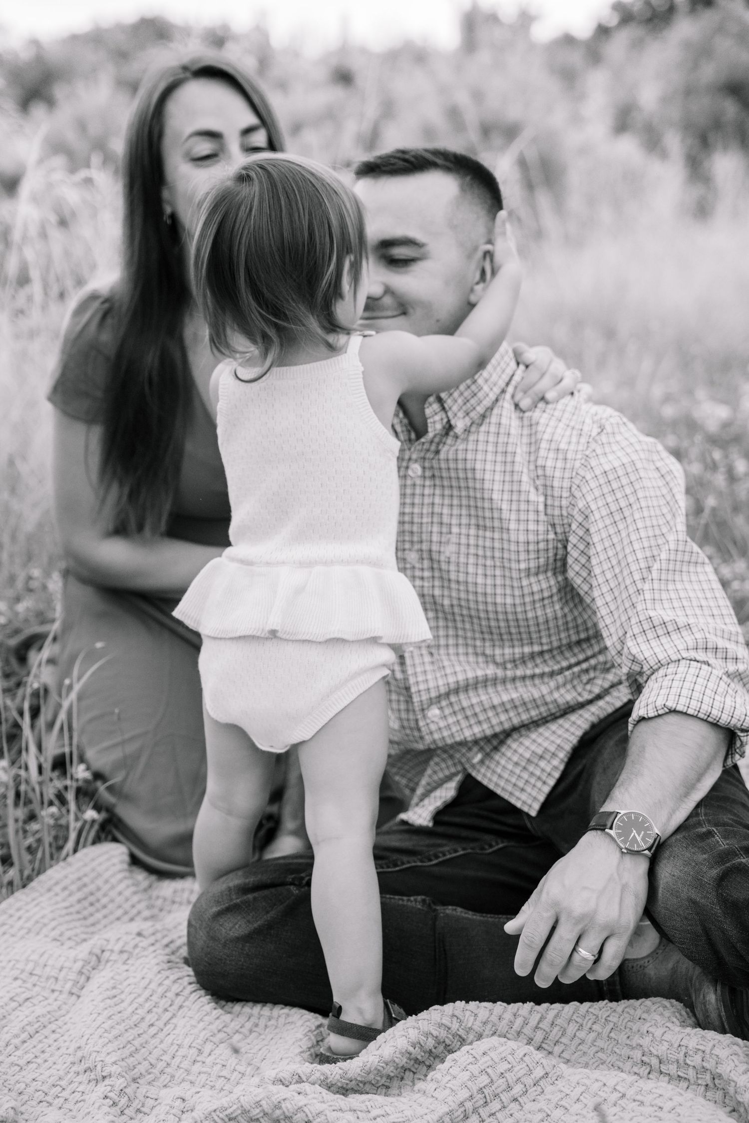 Black and white photo of a young family sitting in a field, with a toddler girl touching her father's face.