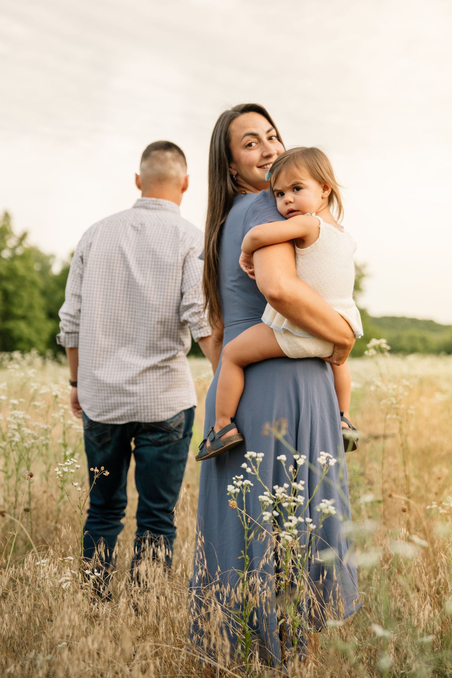 Mother in blue dress holds toddler while father stands in wildflower field at sunset.