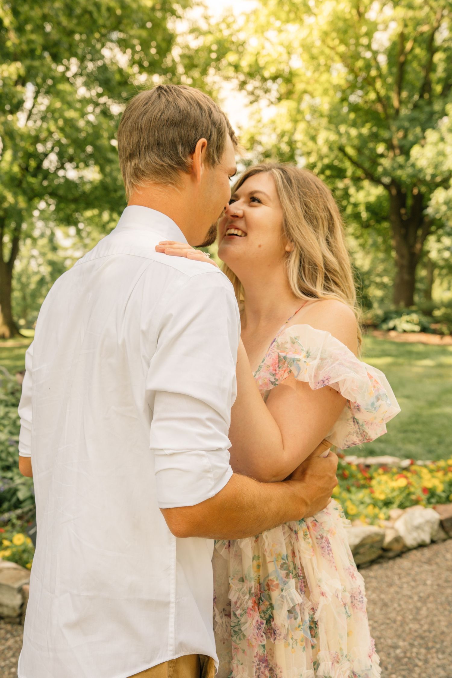 Couple embracing in a lush garden, woman in floral dress smiling up at man in white shirt.