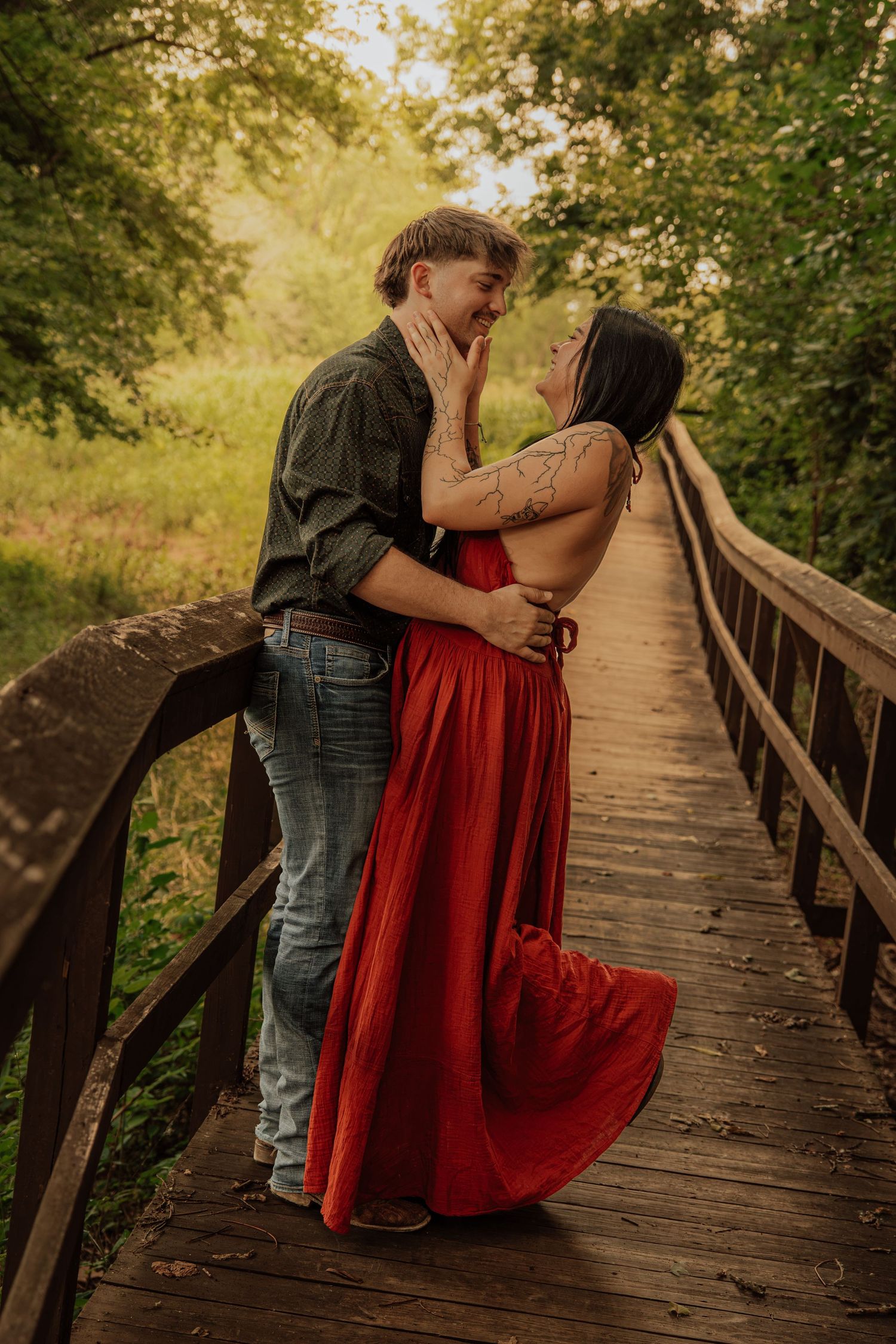 Couple sharing a romantic moment on a wooden bridge surrounded by lush green trees, woman in flowing red dress.
