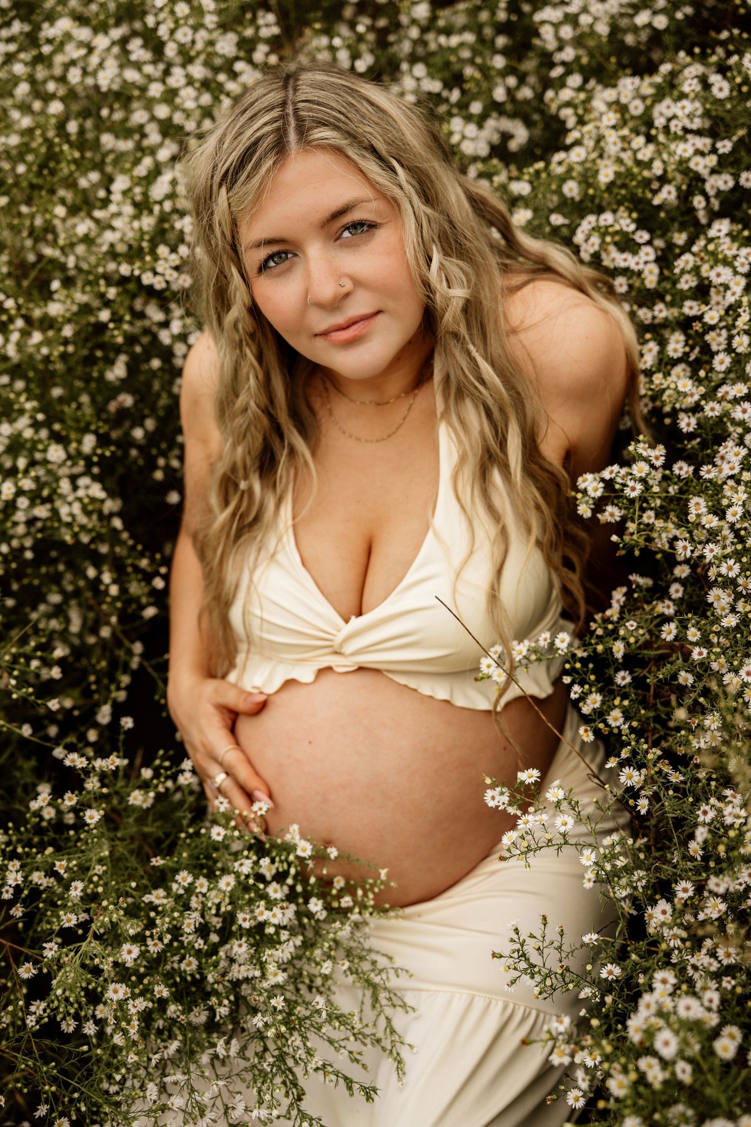 Pregnant woman in cream two-piece outfit surrounded by white wildflowers, gently holding her baby bump.