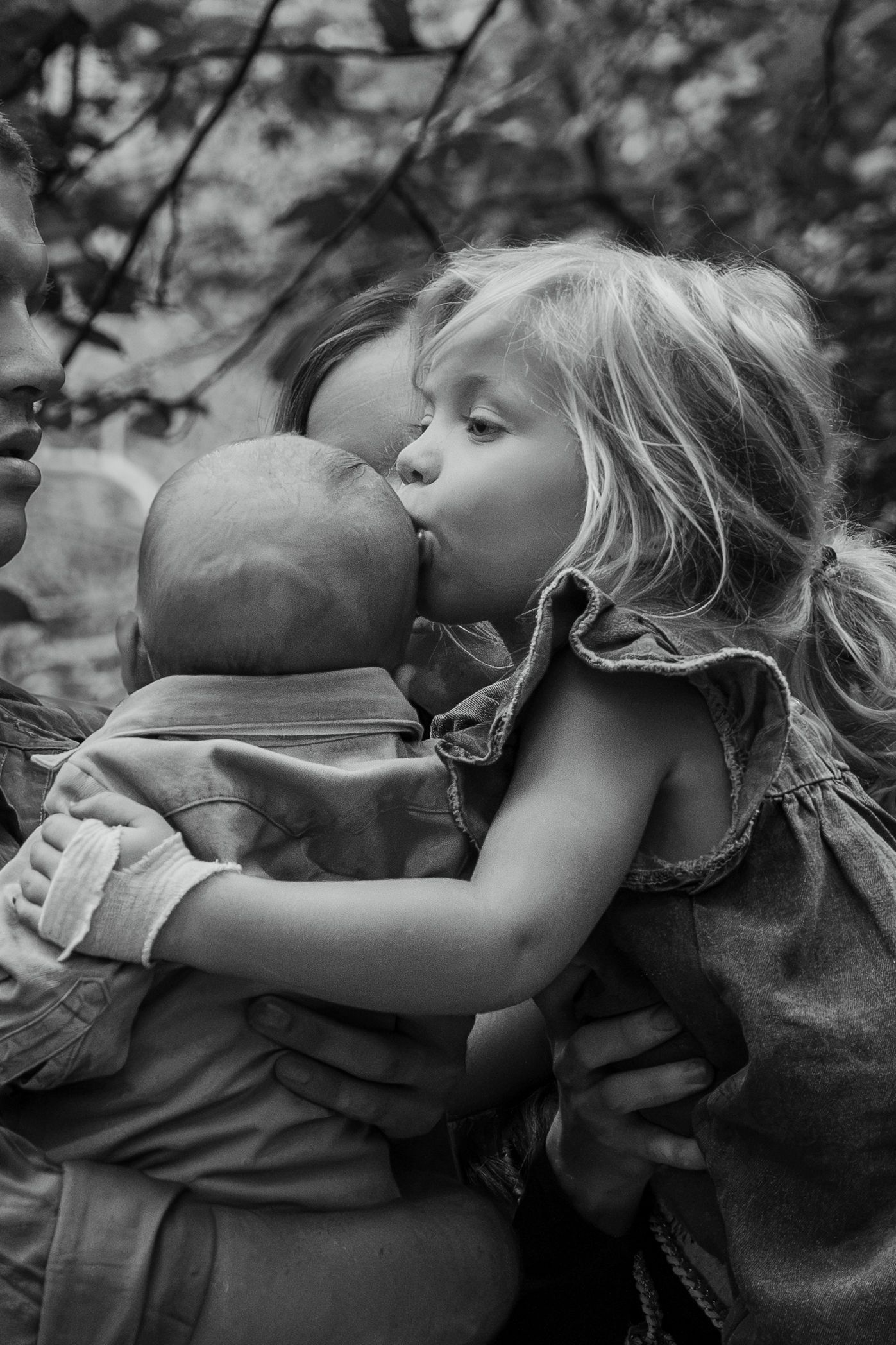 Black and white photo of a young girl kissing a baby sibling while being held by an adult outdoors.