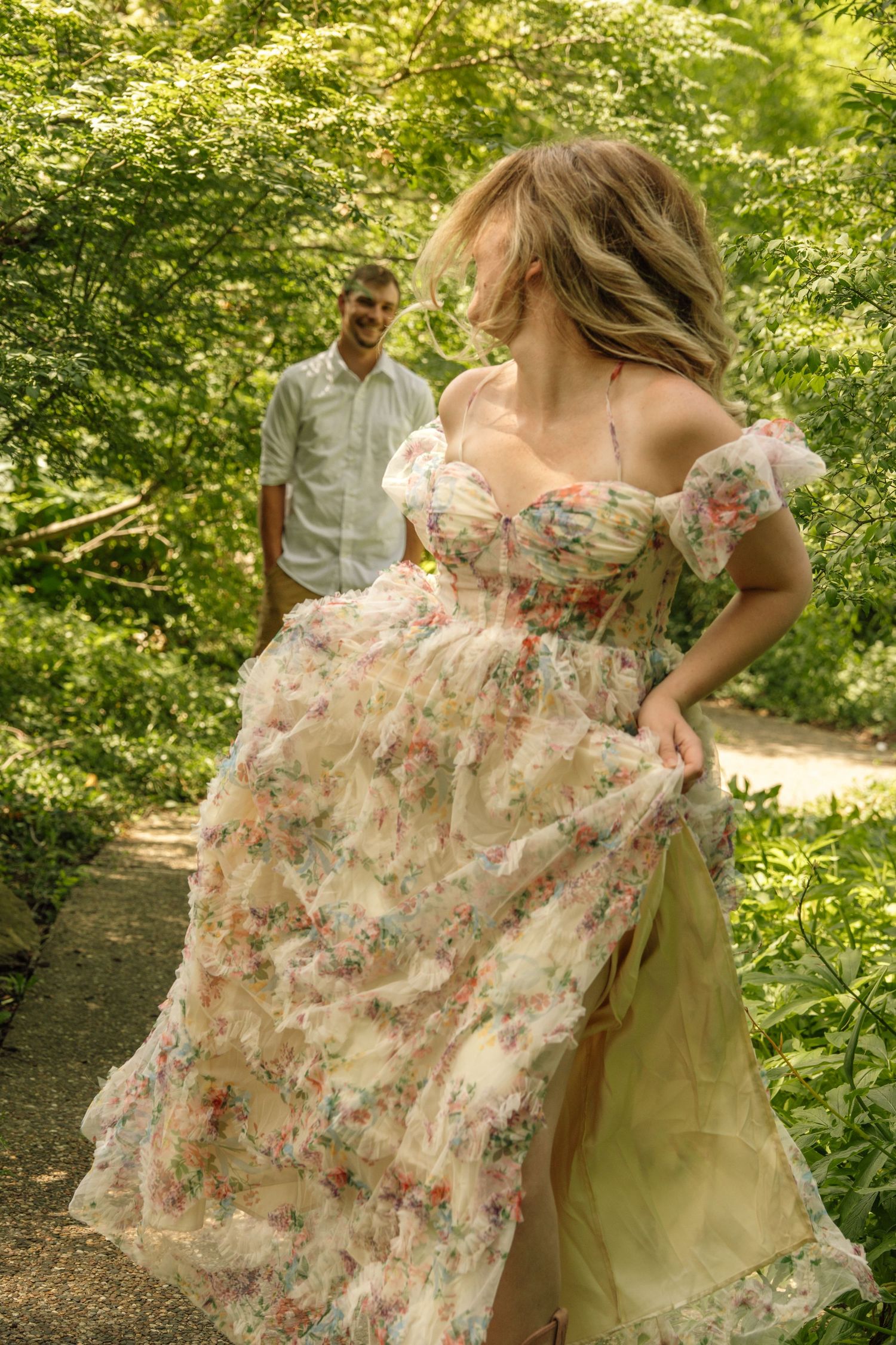 Woman in floral dress twirling on garden path while man smiles in background among lush green trees.