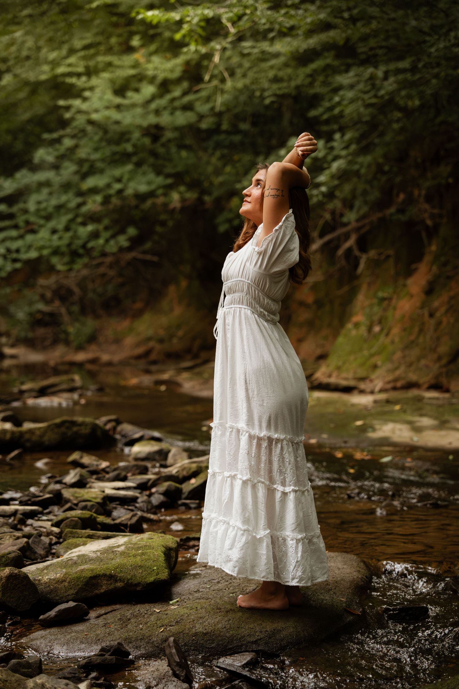Woman in white boho dress standing barefoot on rocks by a forest stream, arms raised, eyes closed in peaceful bliss.