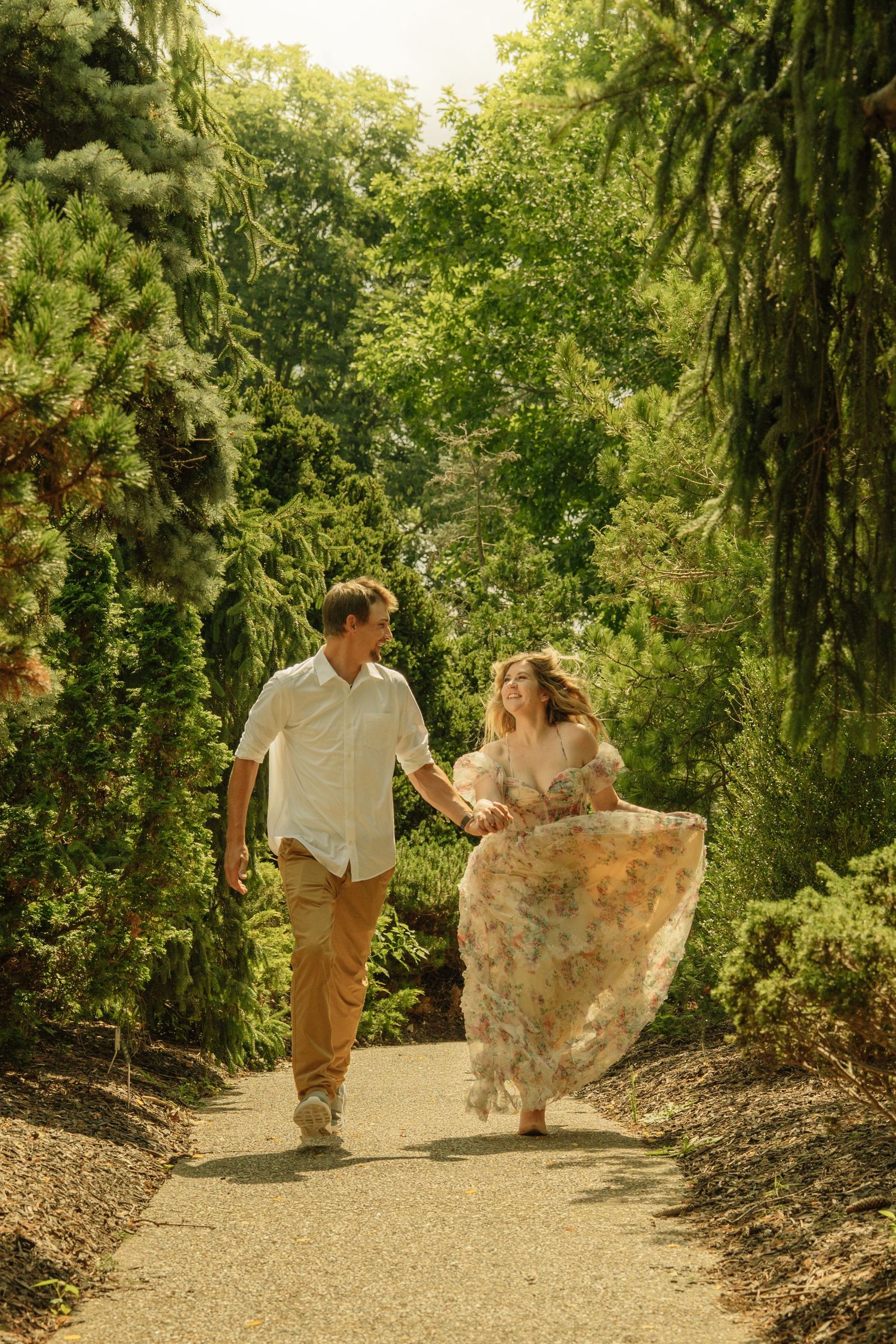 Couple holding hands walking on garden path, woman in floral dress, man in white shirt and tan pants, lush green trees.