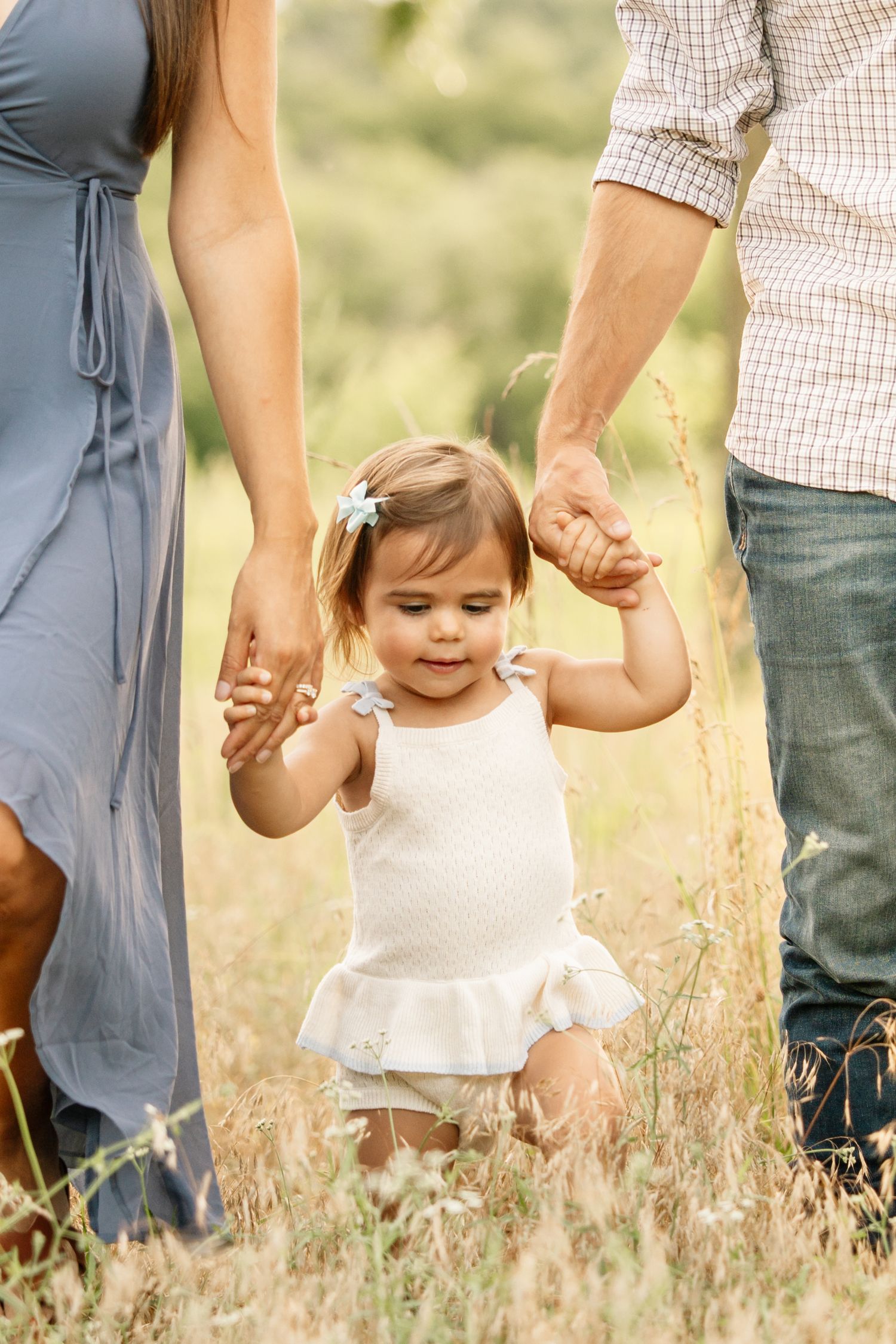 A toddler girl in white dress holds parents hands while learning to walk in a sunny field.