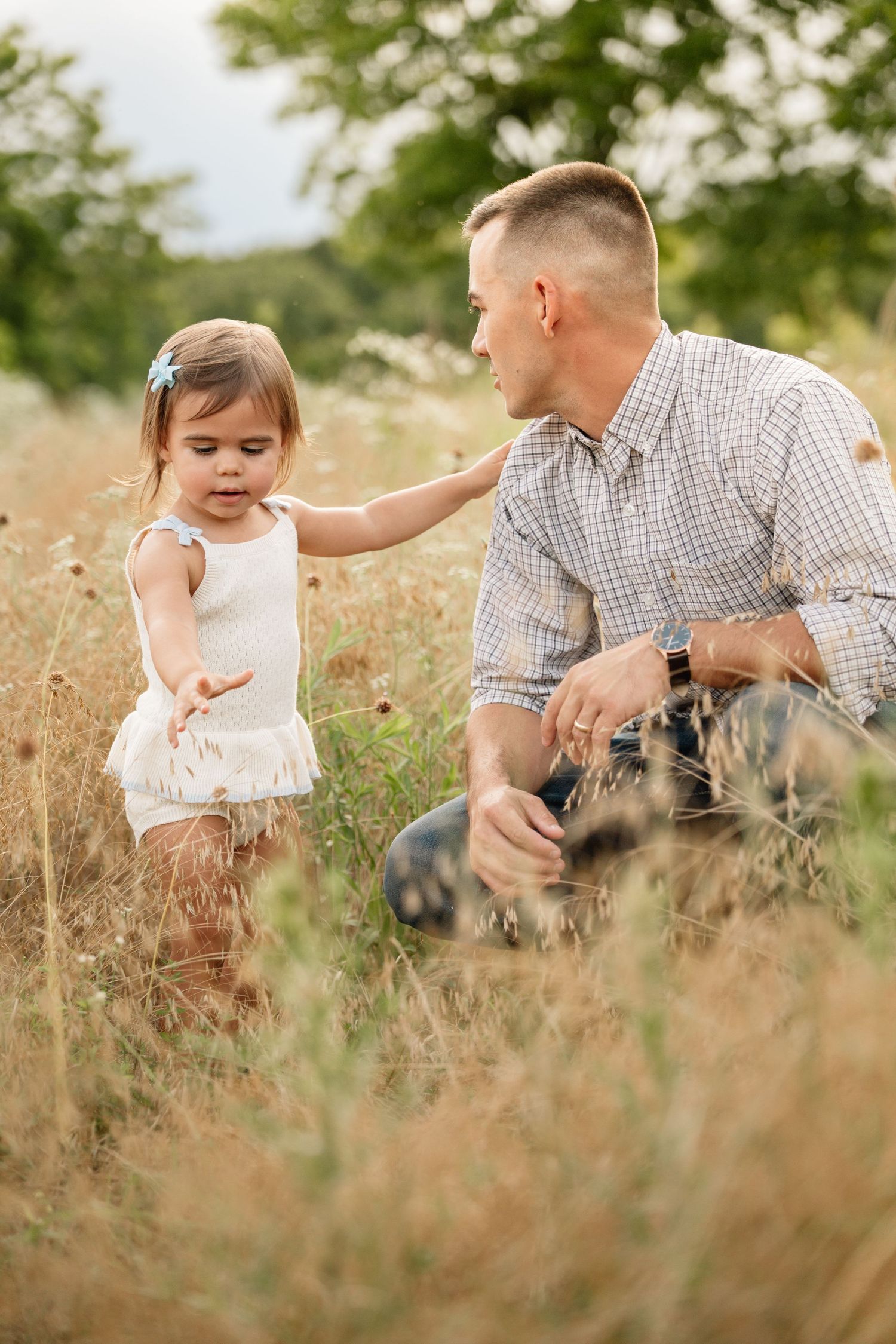 Father crouches in tall grass field as young toddler daughter reaches toward him.