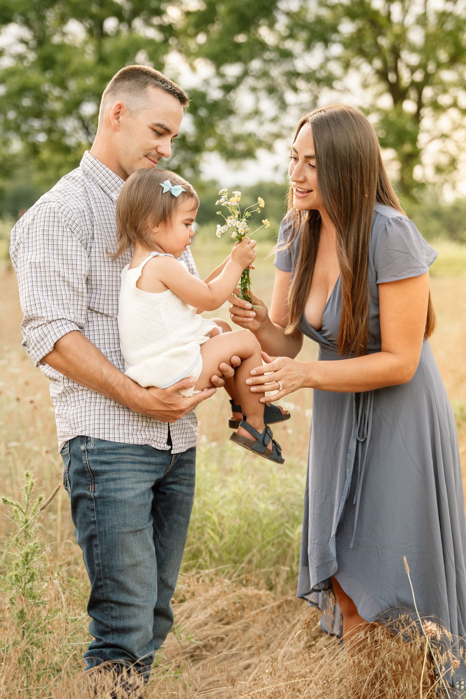 A happy young family with a baby girl holding wildflowers in a sunny field, dad in plaid shirt and mom in gray dress.