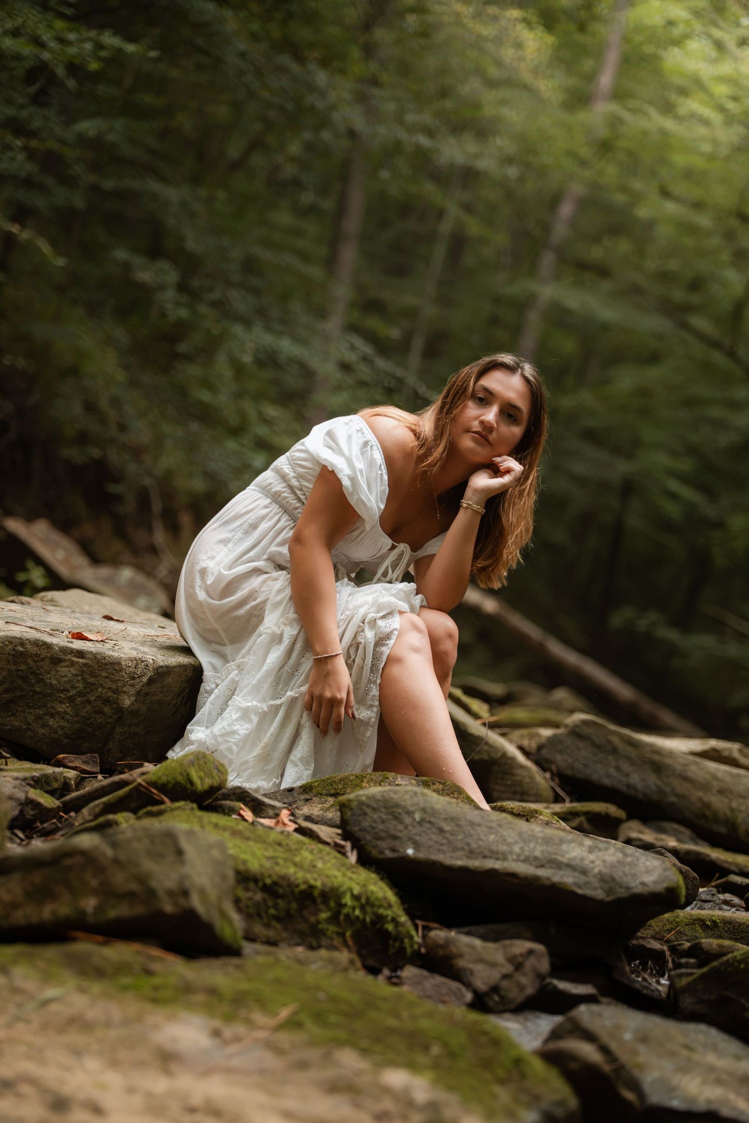 Woman in white dress sitting on moss-covered rocks in a sunlit forest setting.