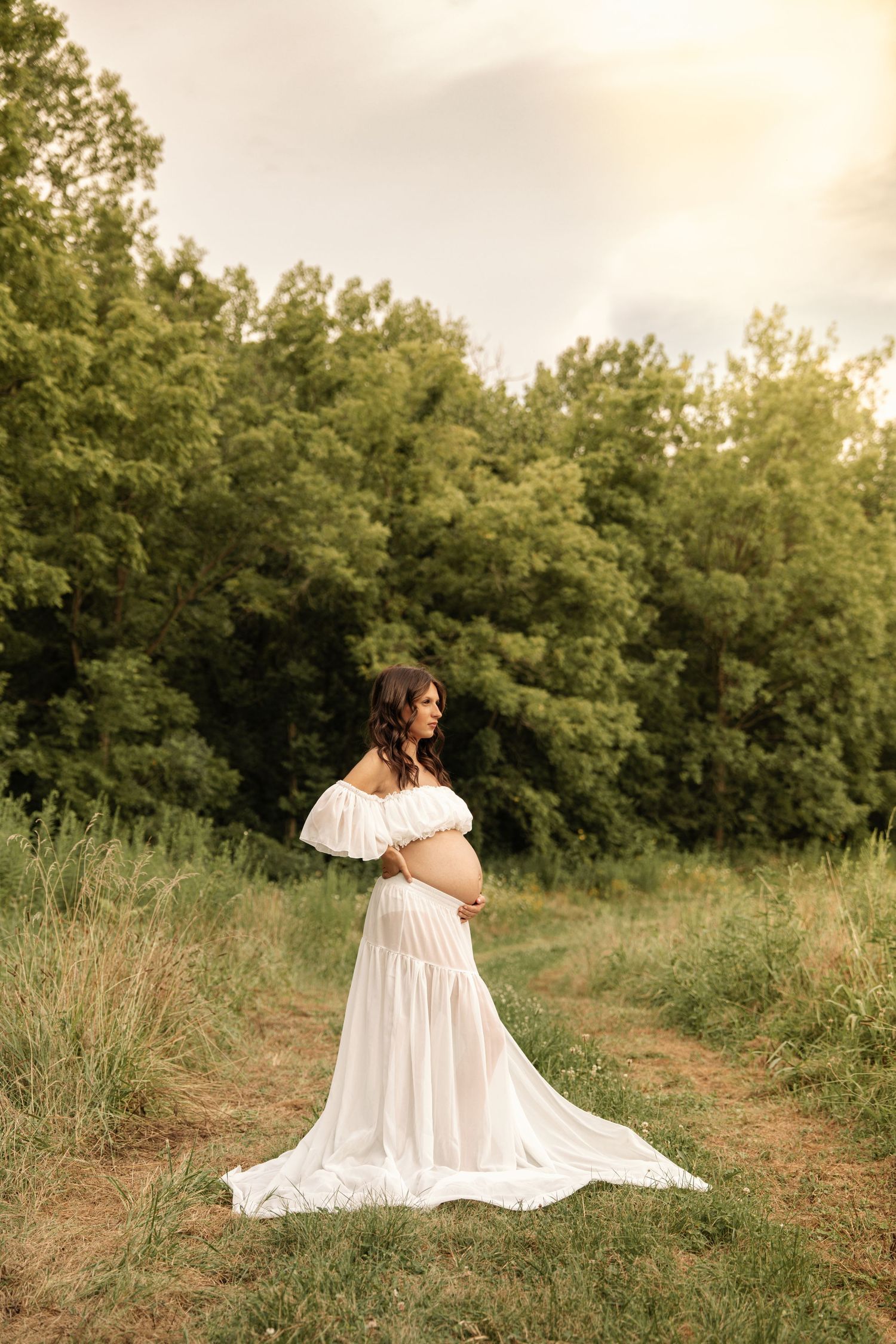 Pregnant woman in white off-shoulder gown cradling belly in a sunlit green meadow path.