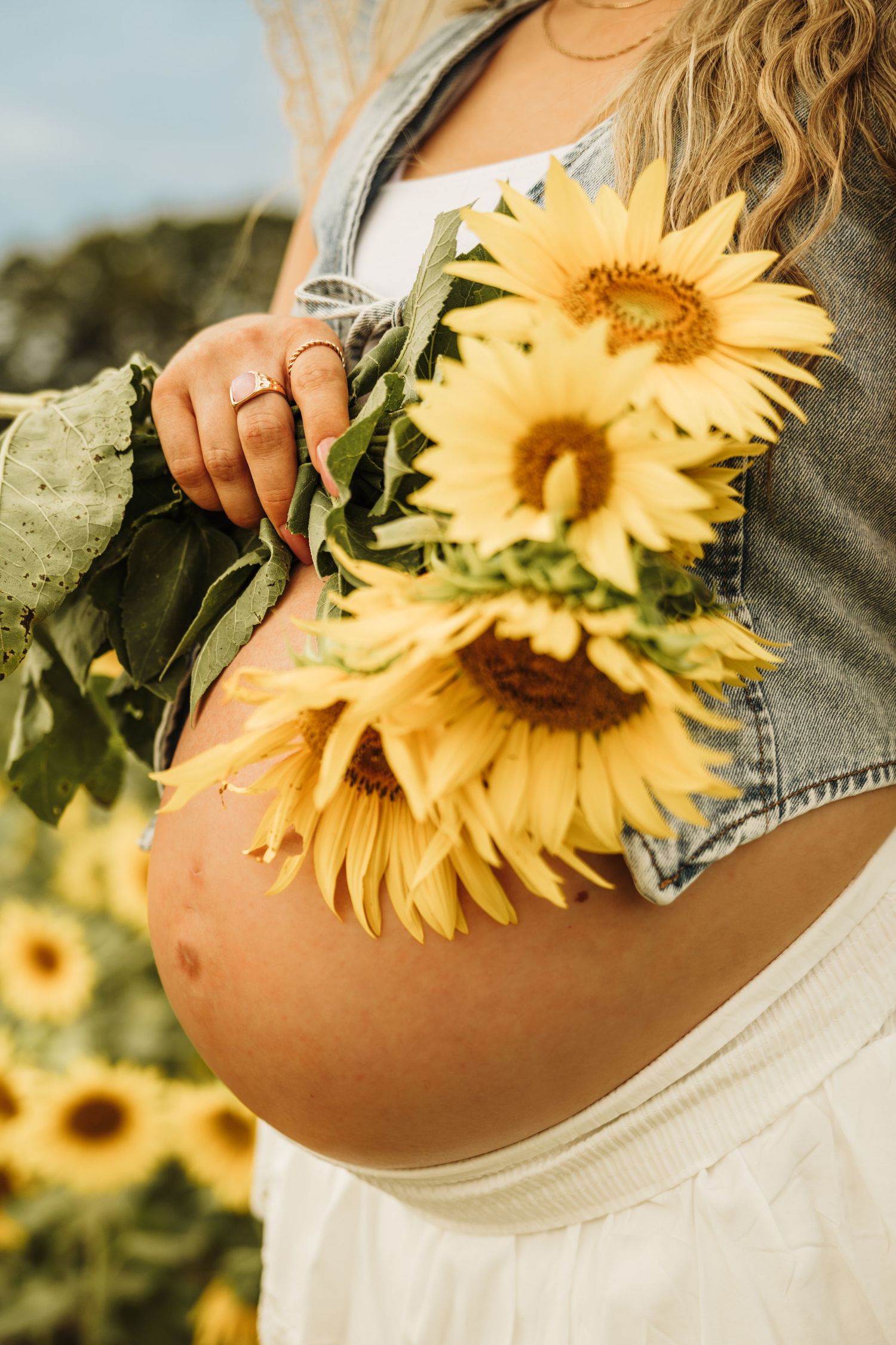 Pregnant woman holding sunflowers in a sunflower field, wearing a denim vest and white skirt.