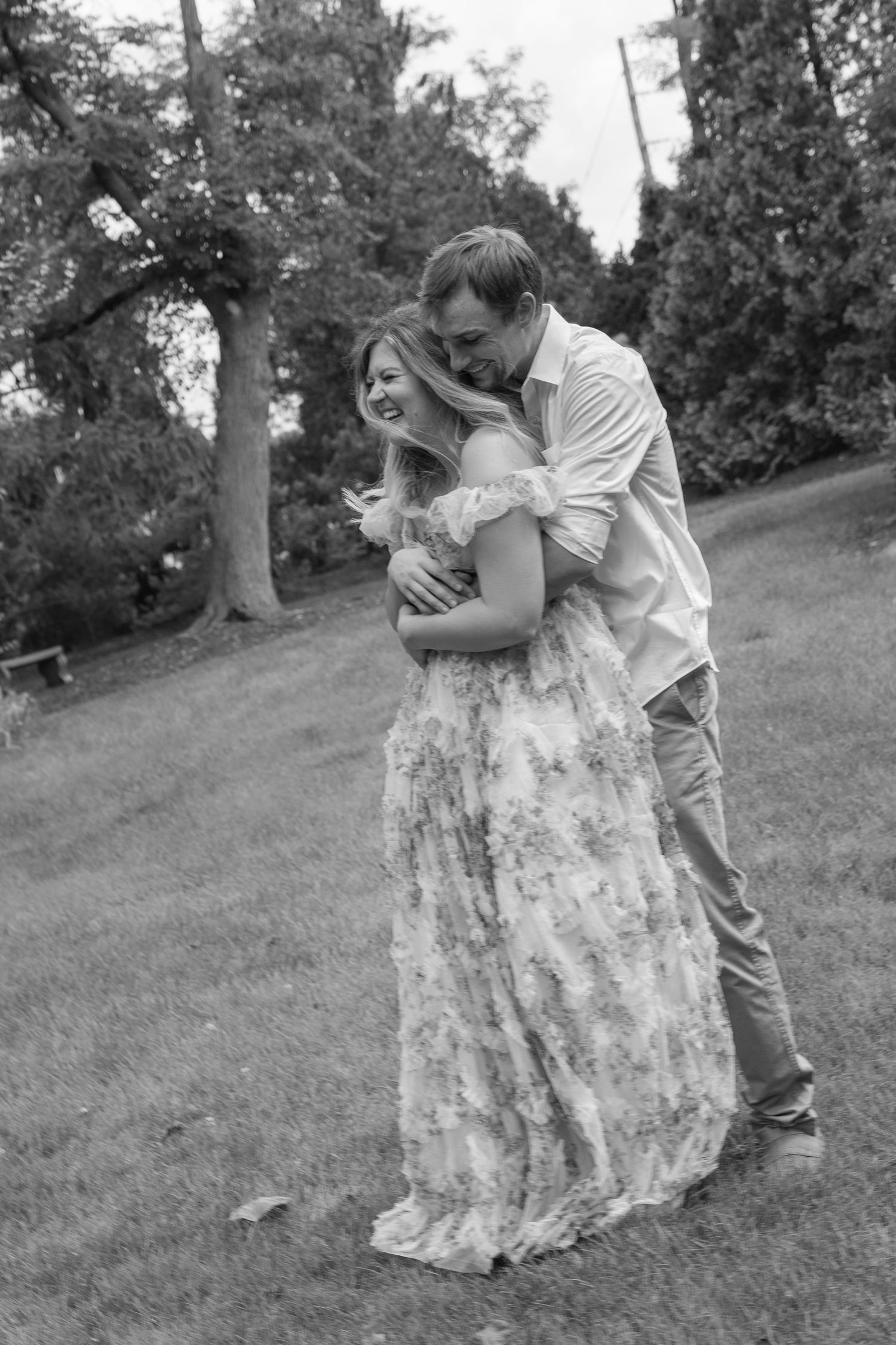 Black and white photo of a joyful couple hugging and laughing together on a grassy park lawn.