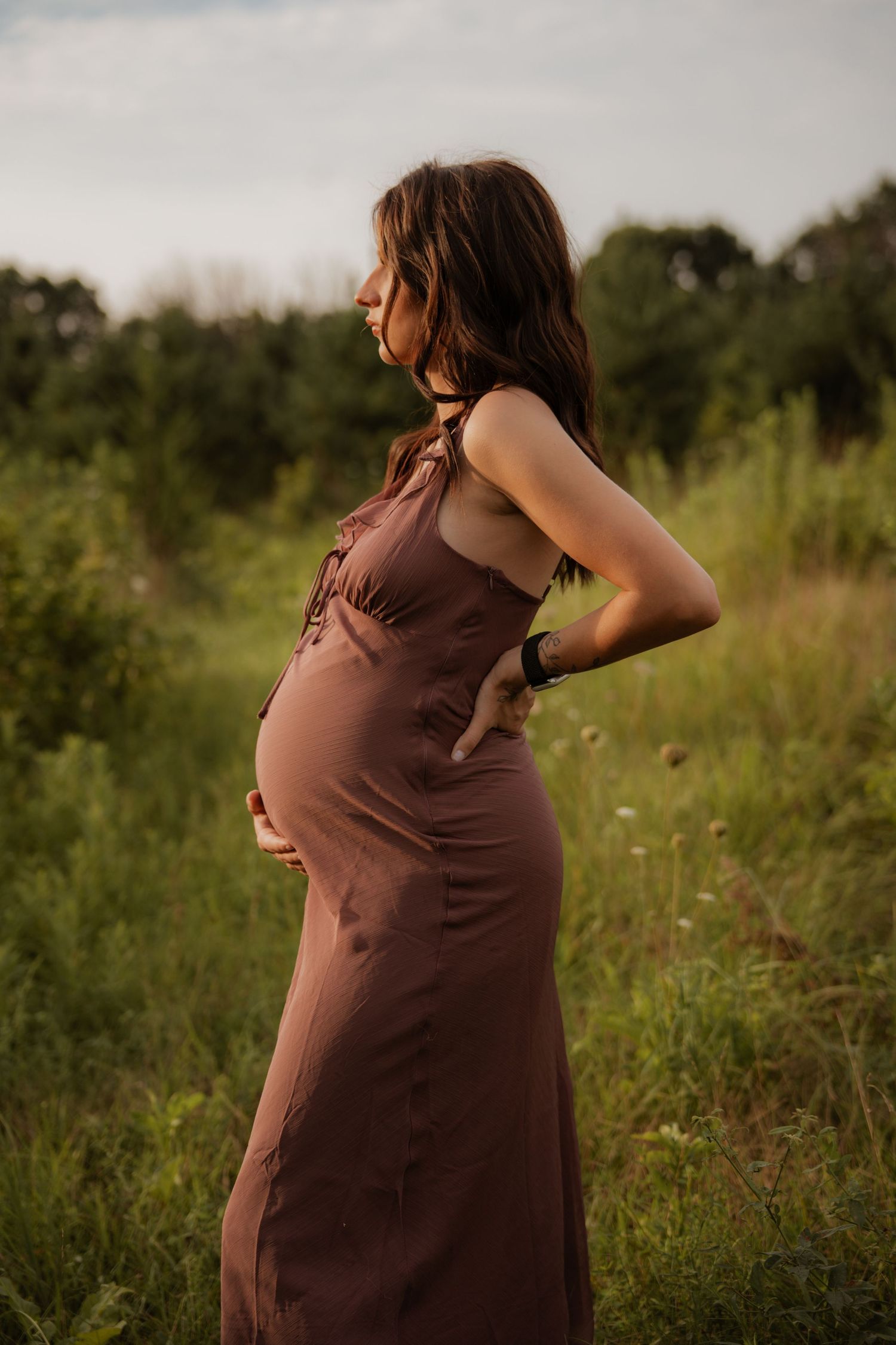Side view of pregnant woman in brown halter maxi dress standing in lush green field at golden hour.