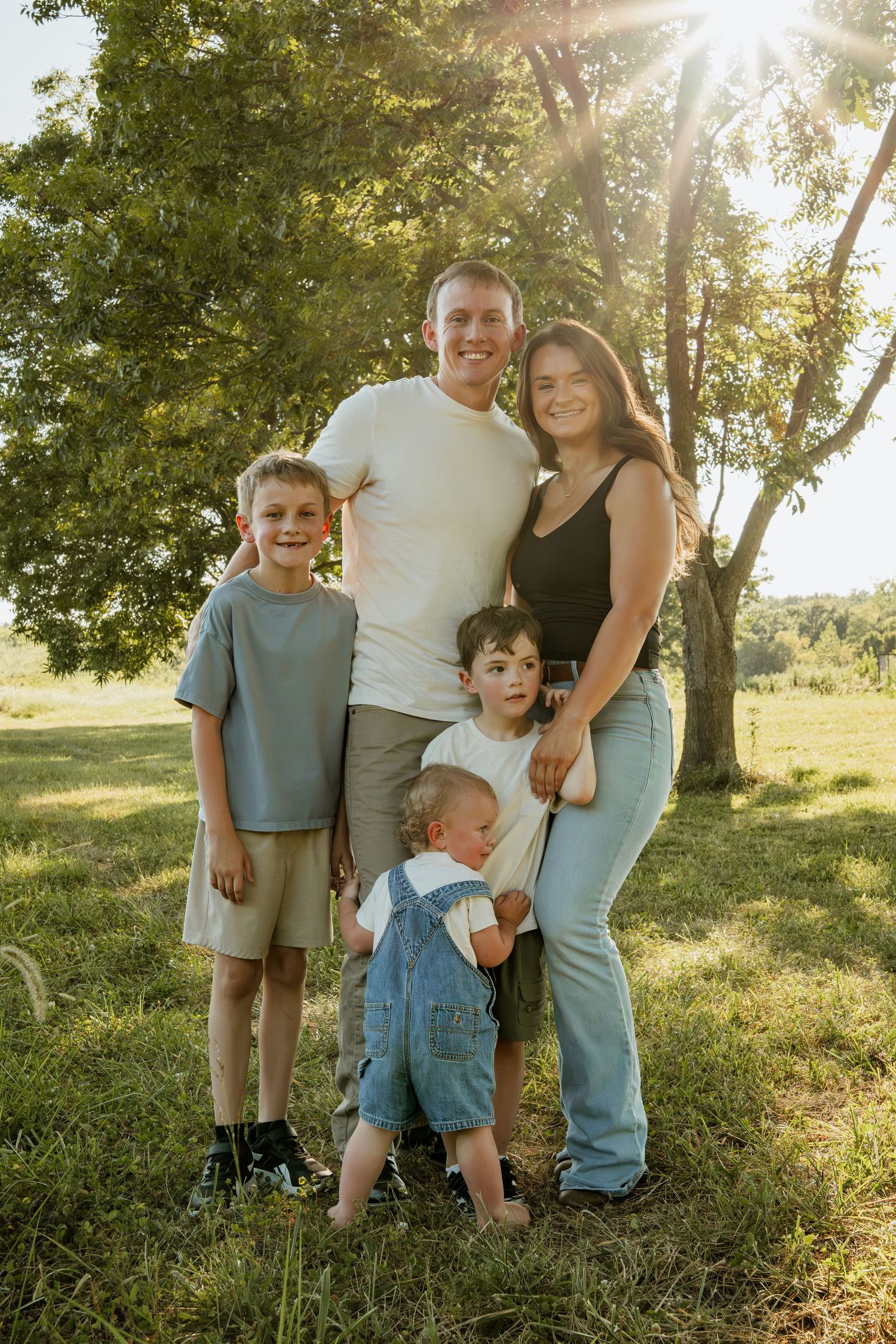 Happy family of five posing outdoors in golden sunlight with lush green trees in background.