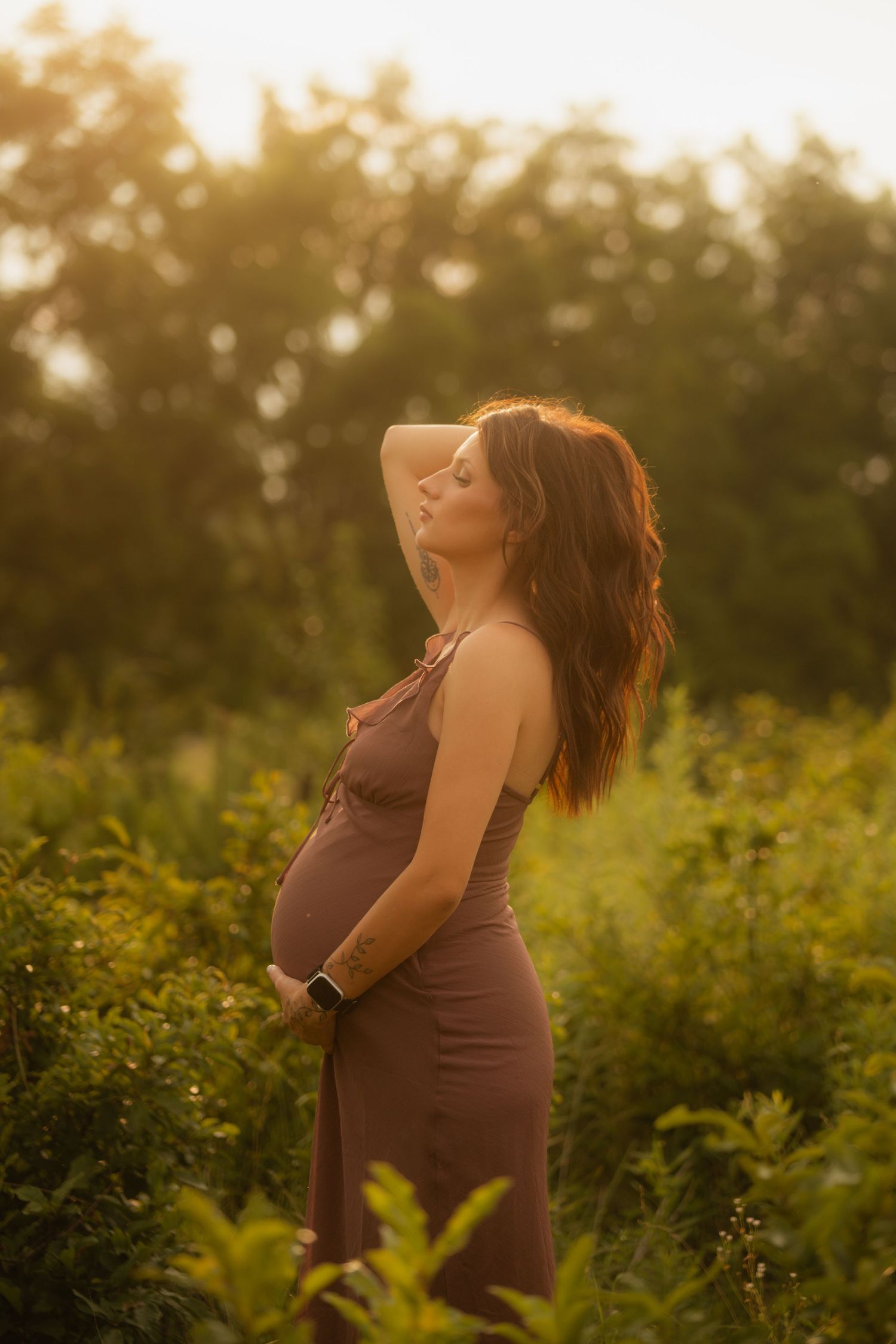Glowing pregnant woman in brown dress cradles bump with eyes closed amid lush greenery in golden sunset.