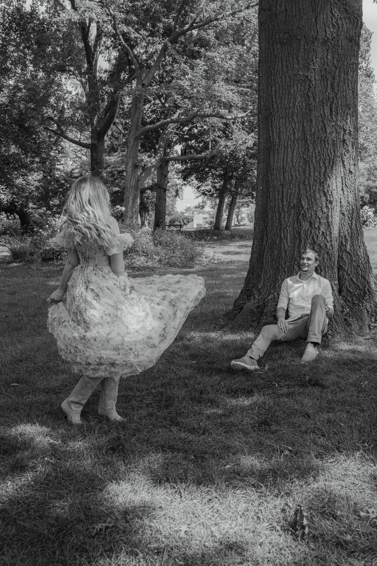 Black and white photo of woman twirling in floral dress as man watches smiling against a large tree.