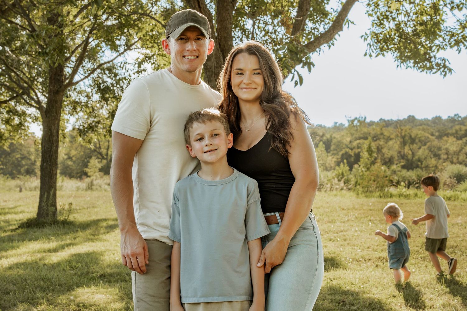 Family of three posing outdoors in a sunny green park, with a child playing in the background.