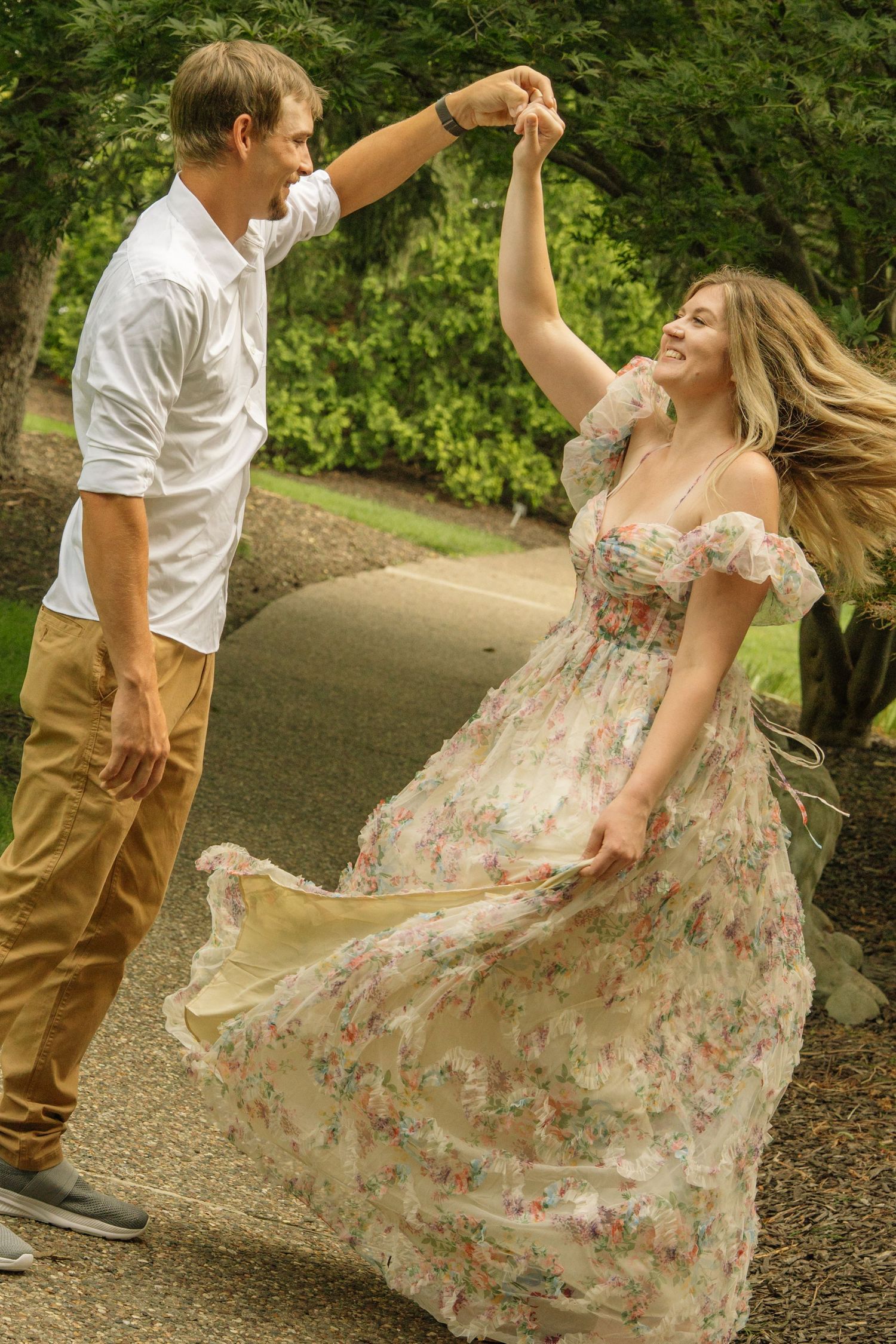 Couple dancing outdoors in a park, woman in floral dress spinning joyfully while man in white shirt leads.