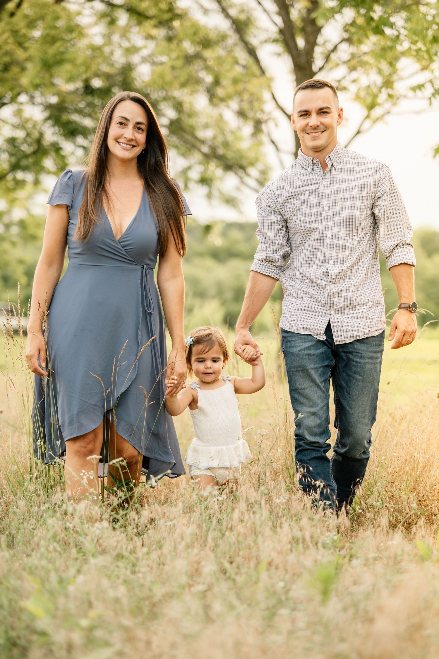 Happy family with toddler walking through a sunny meadow, parents holding child's hands.