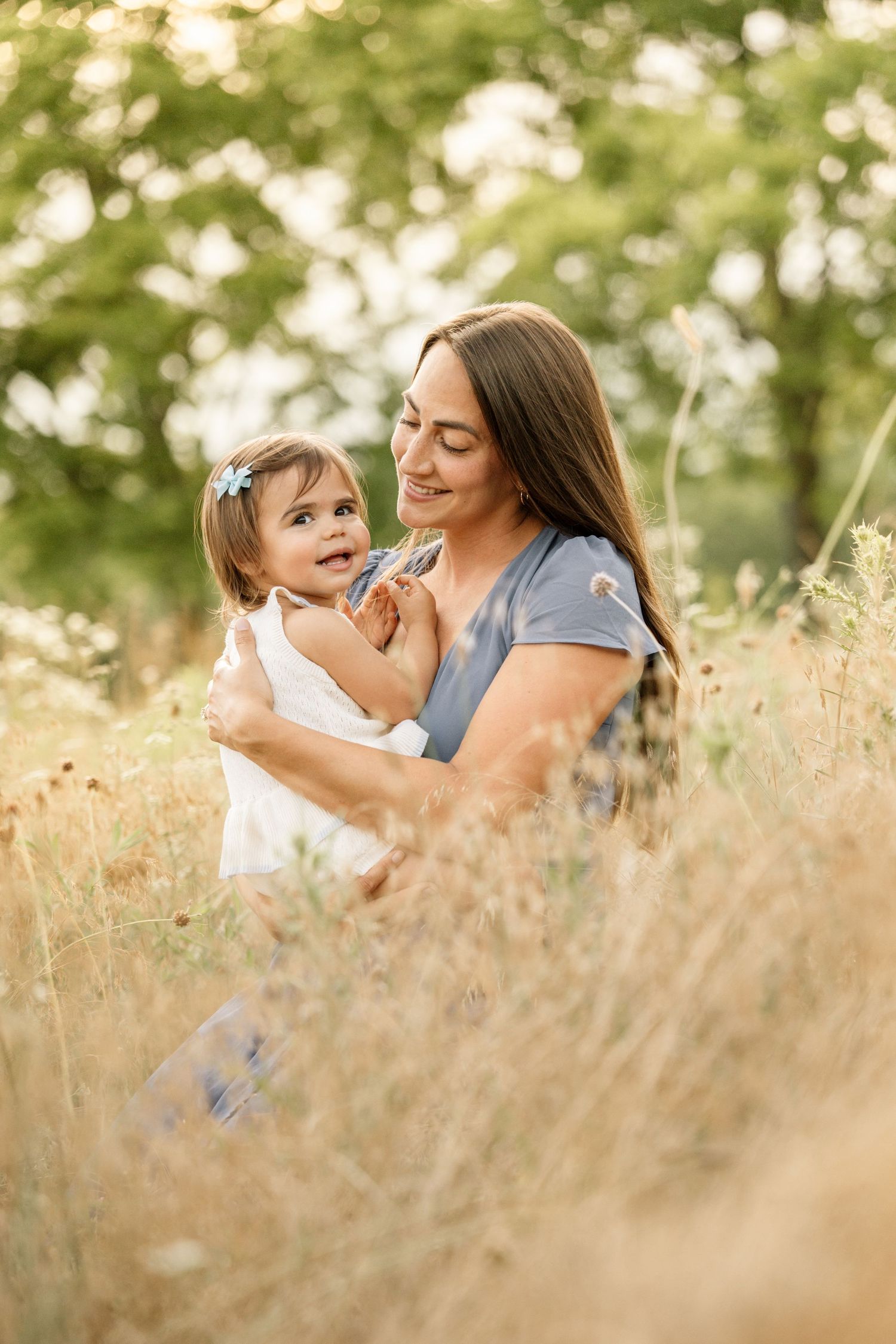 A smiling mother sits in a golden meadow holding her happy toddler daughter in warm sunlight.