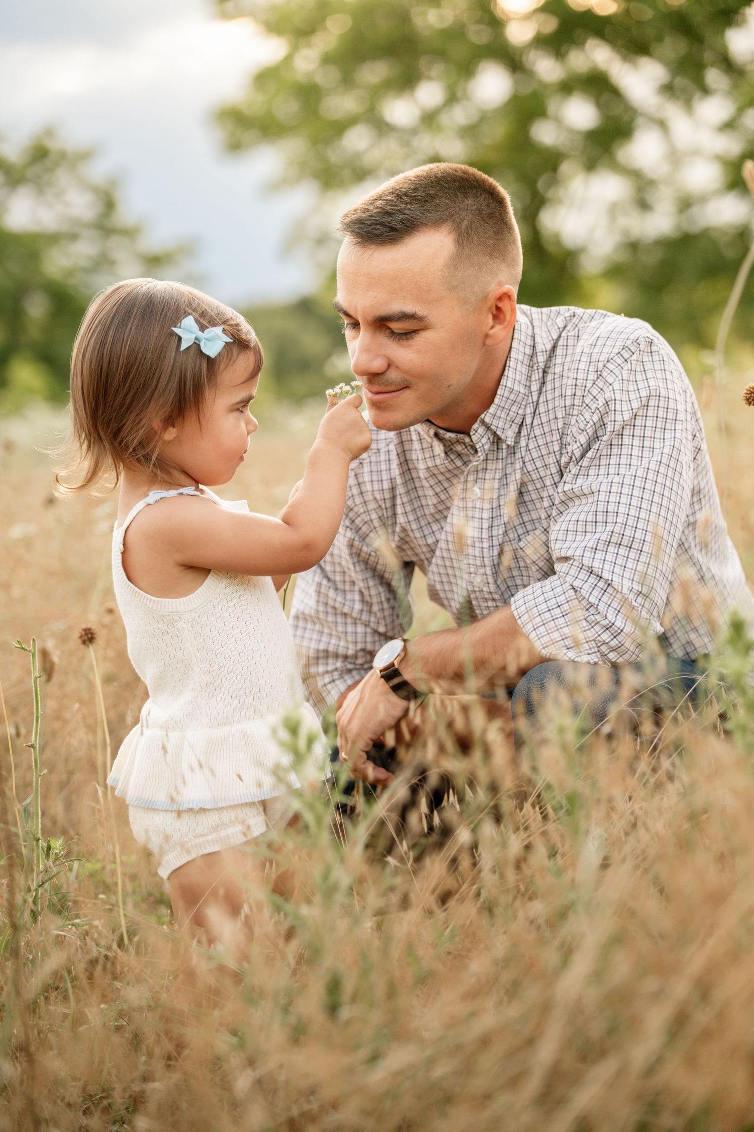 A smiling father crouches in a golden field as his toddler daughter touches his face with a flower.