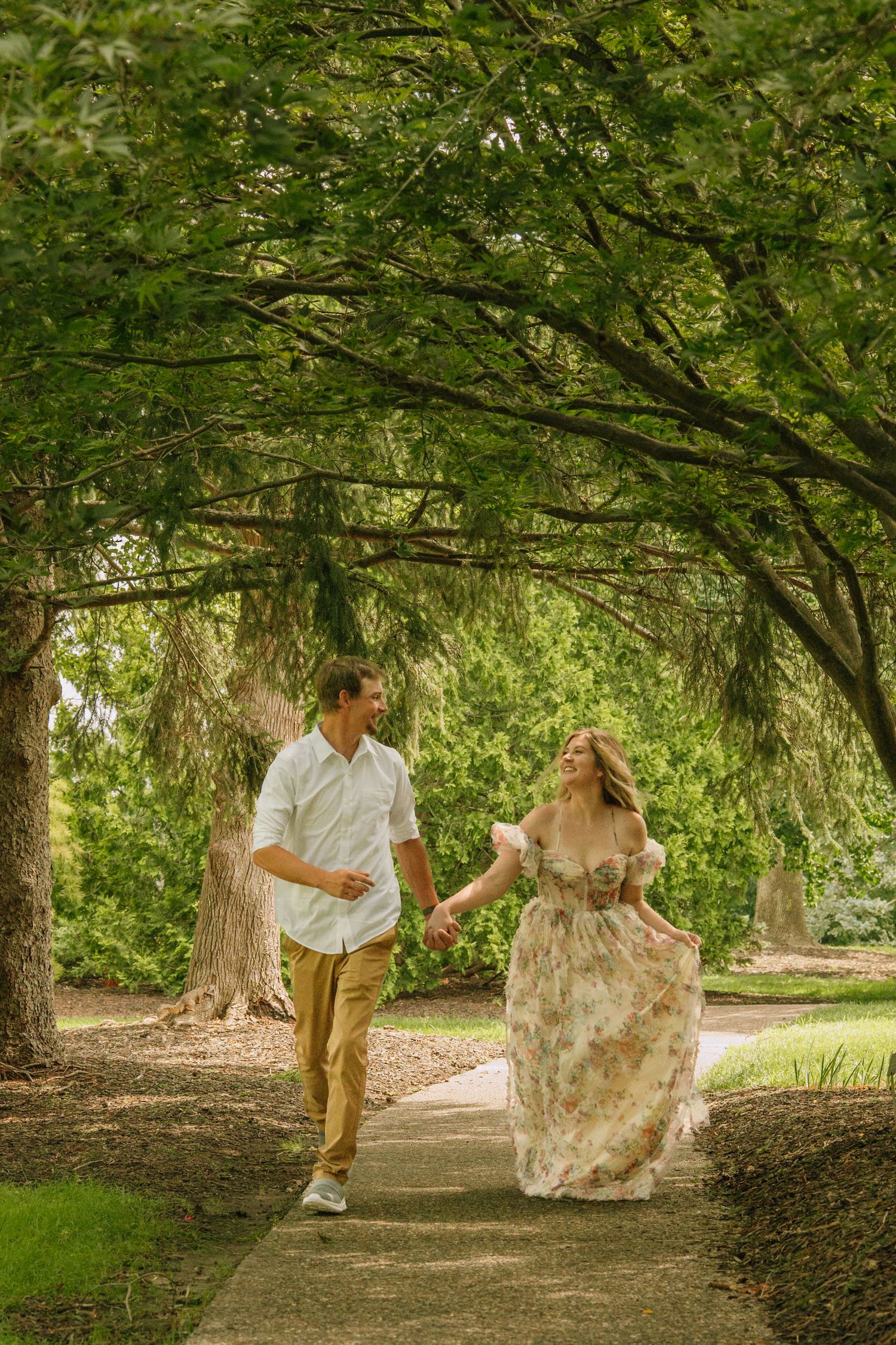 Couple holding hands walking along a tree-lined garden path, woman in floral dress, man in white shirt.