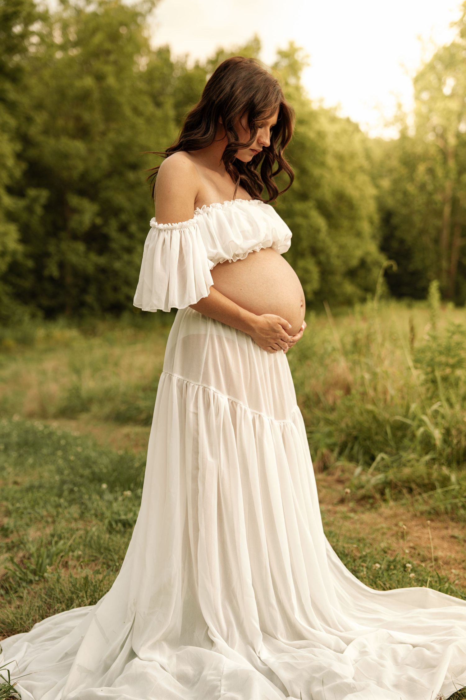 Pregnant woman in white off-shoulder gown cradling baby bump outdoors at golden hour.