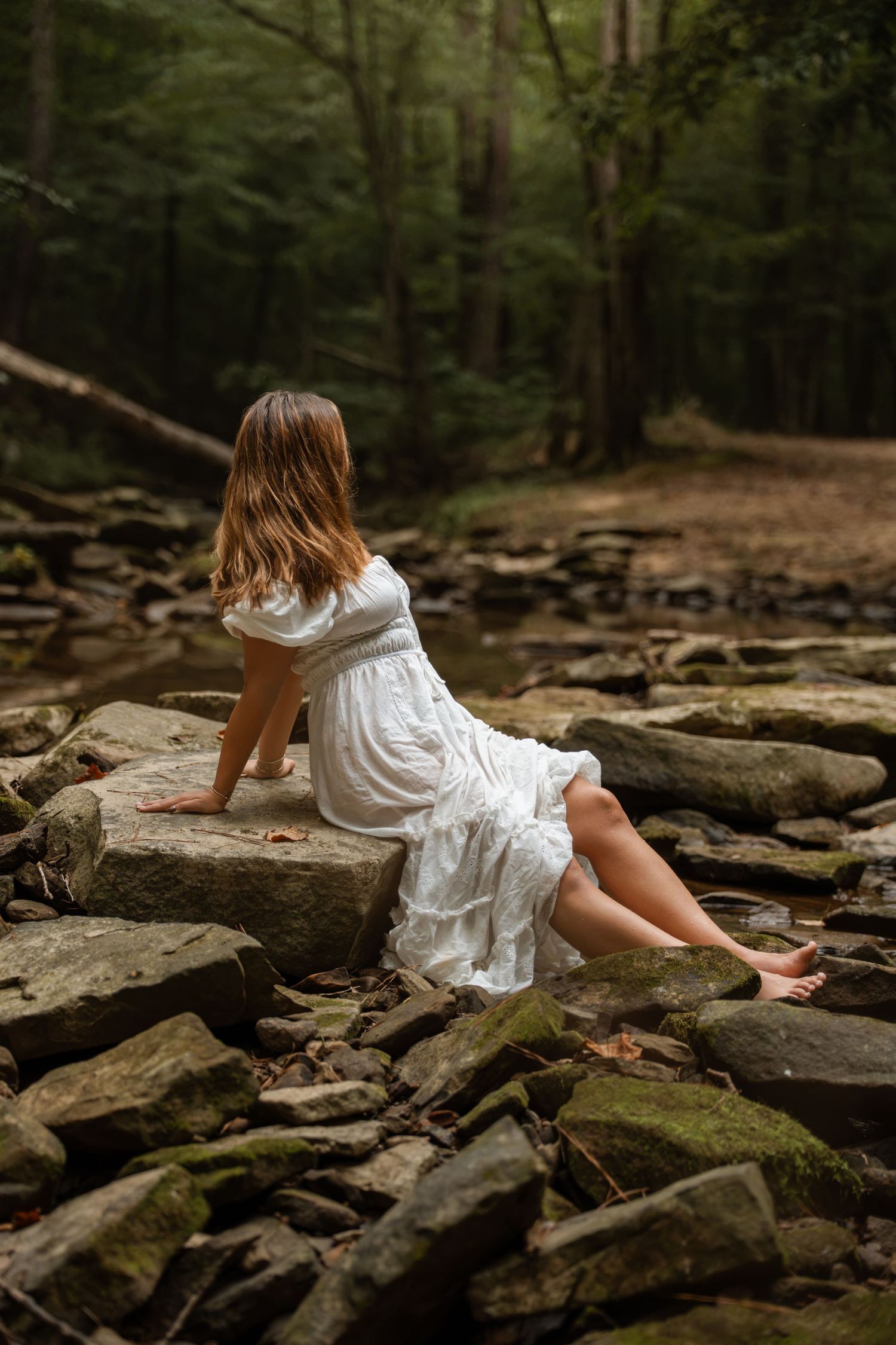 Woman in white dress sitting on rocks with her back turned in a dark forest.