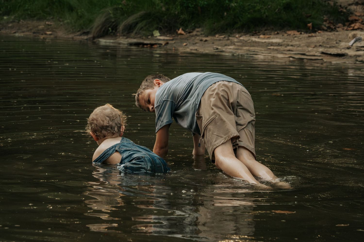 Two children playing and exploring in a shallow river, wading through calm water surrounded by nature.