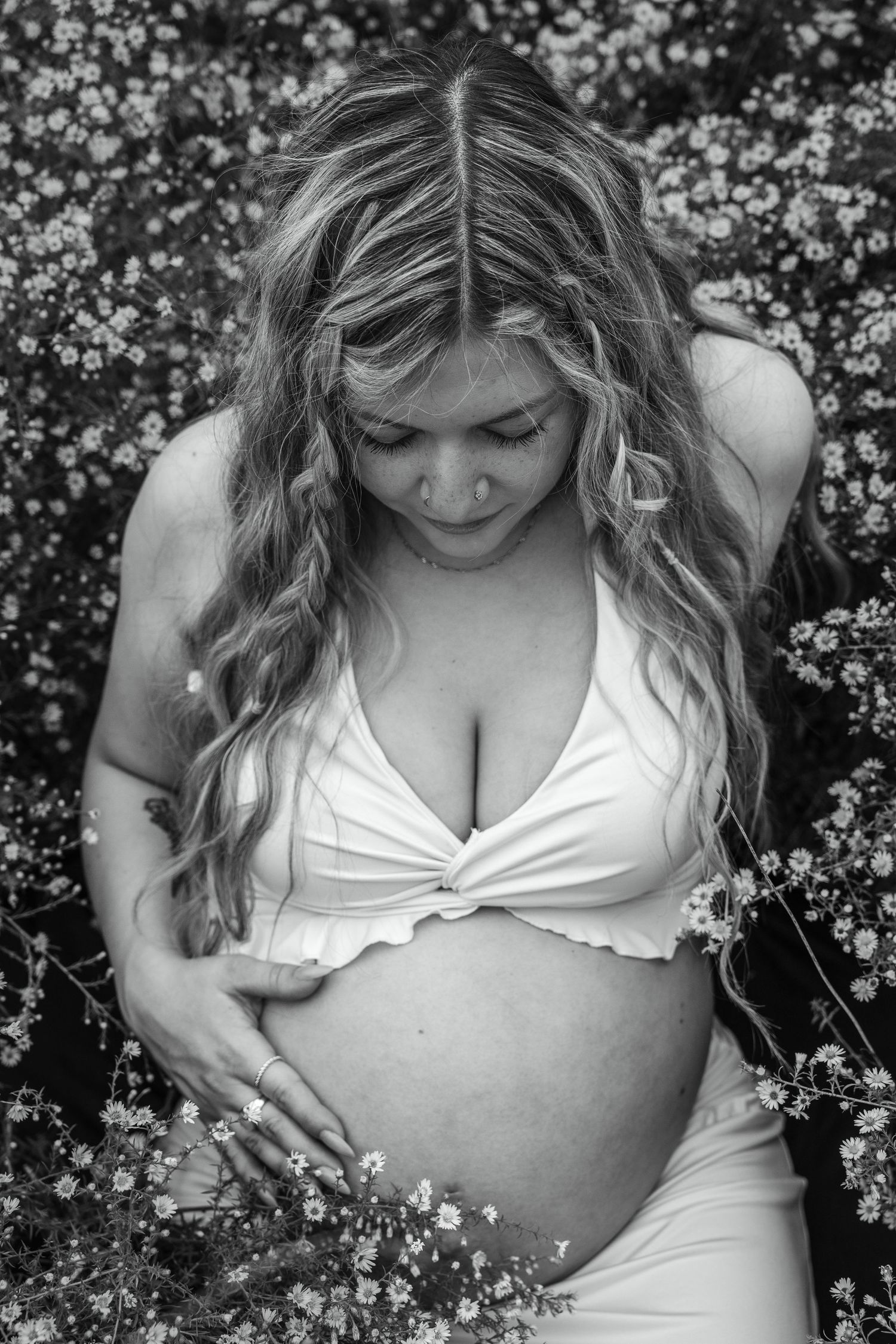 Black and white photo of a pregnant woman in a white crop top cradling her baby bump among wildflowers.