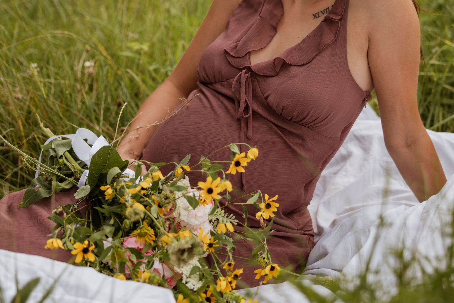Pregnant woman in brown dress sitting in green grass field holding yellow wildflowers bouquet during outdoor maternity photoshoot.