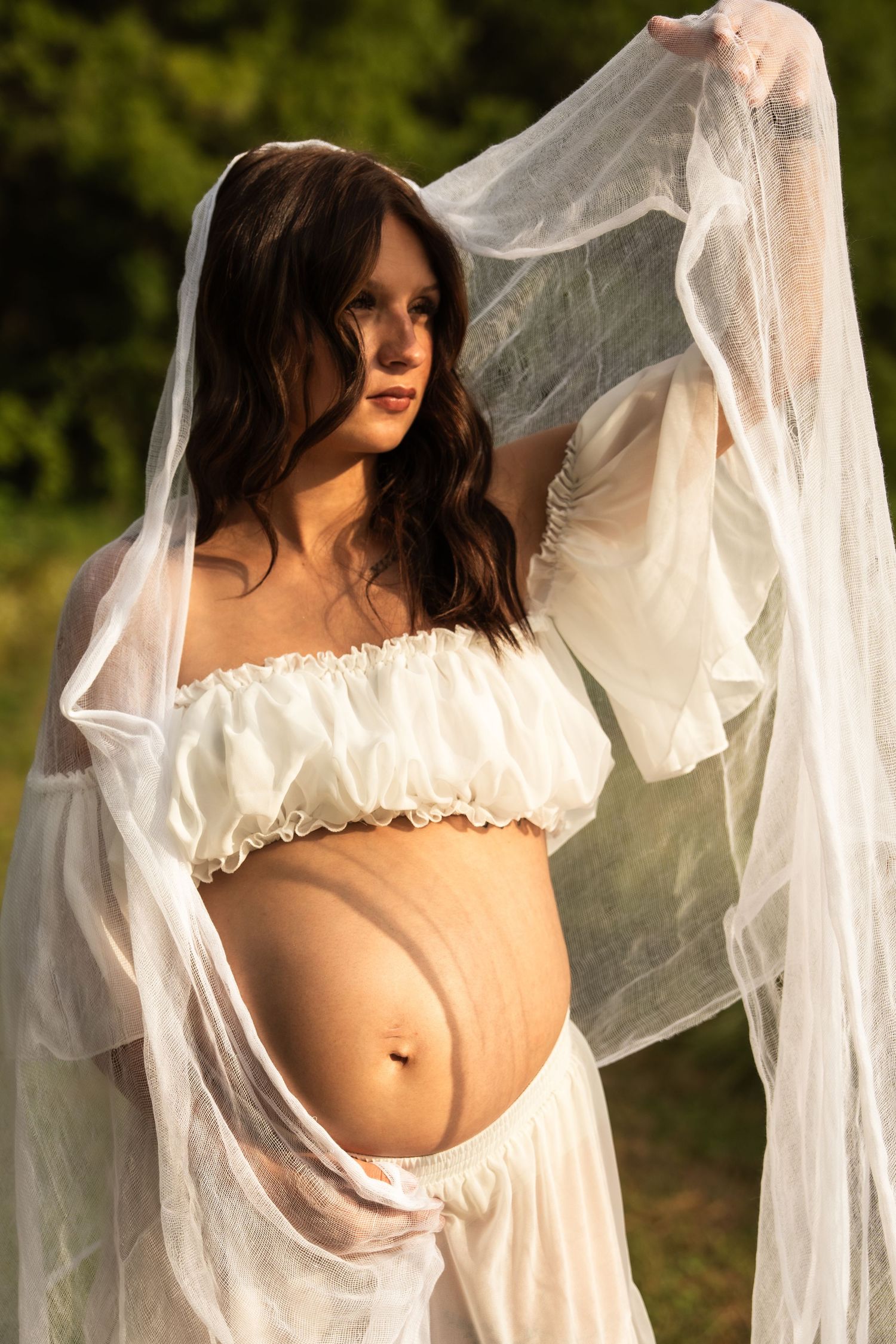 Pregnant woman in white crop top holds sheer fabric overhead, showcasing baby bump in natural light.
